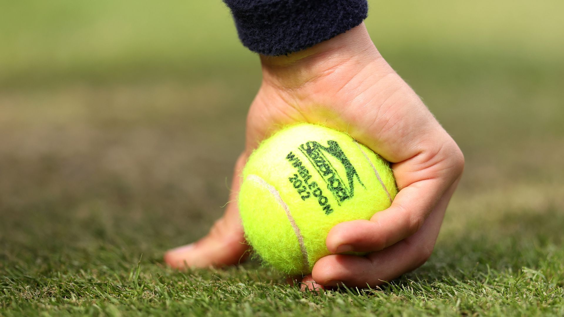 A hand picks up a tennis ball that reads "Wimbledon 2022."