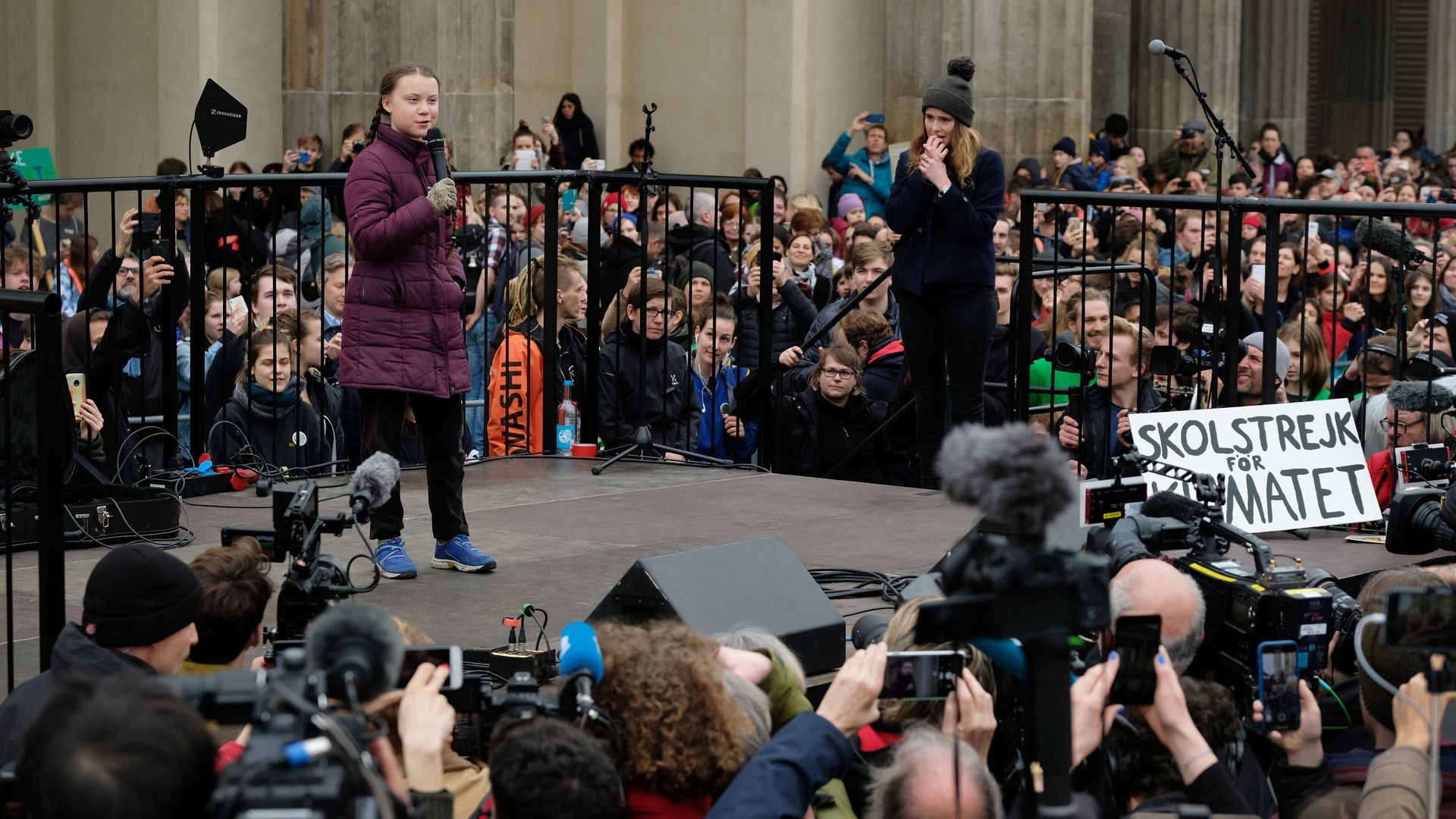 In this image, Greta stands and speaks into a microphone on a small outdoor stage surrounded by a crowd. 