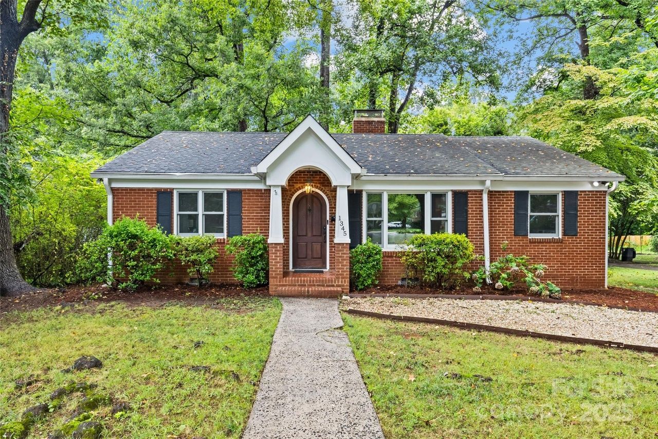 Single-story red brick house with dark shingled roof, arched wooden front door, white trim, black shutters, surrounded by green bushes and tall trees under blue sky.