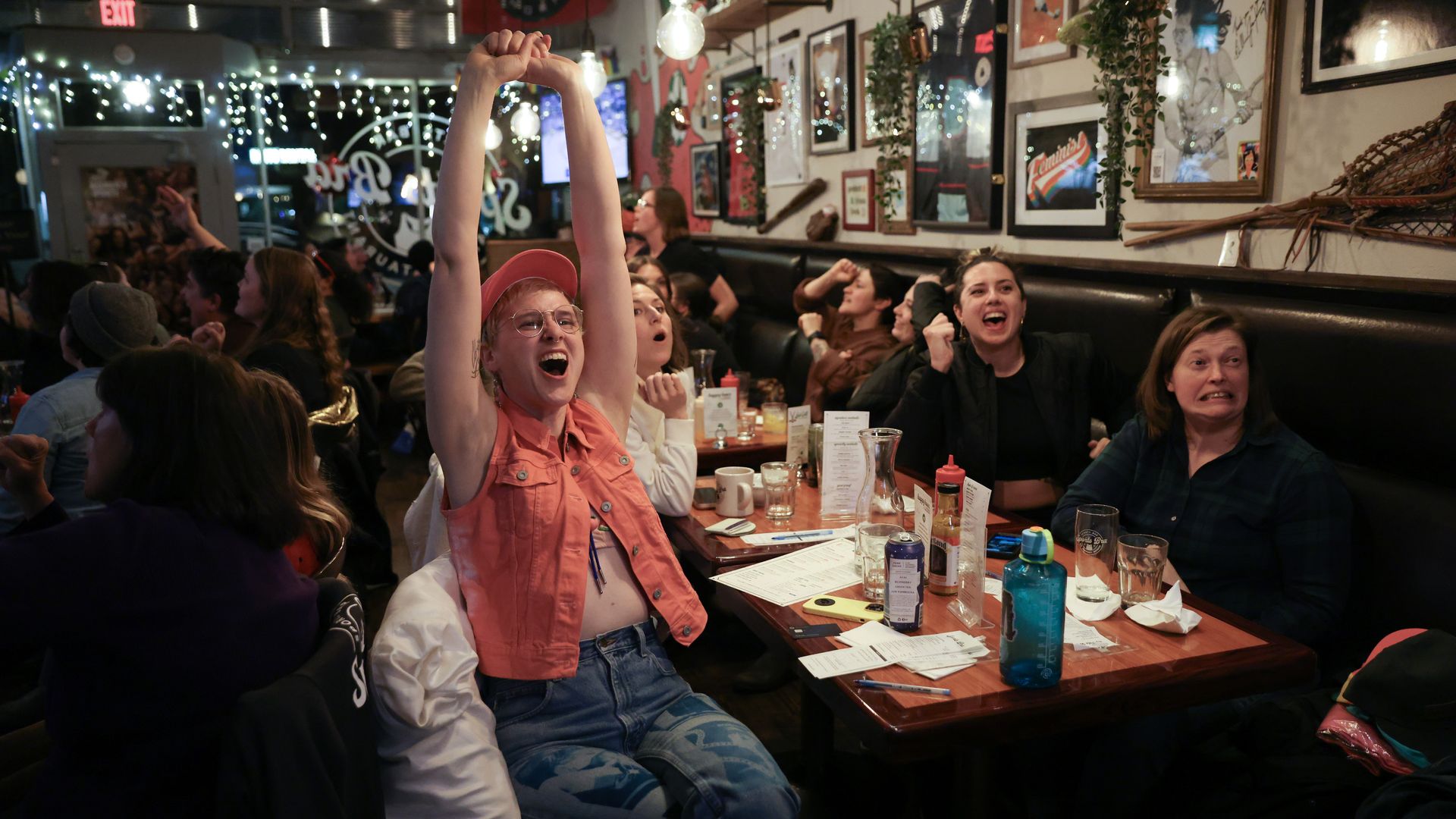 A lively restaurant scene with a woman in an orange shirt and pink cap raising her arms in celebration. A cheering crowd at tables under string lights and framed wall art.