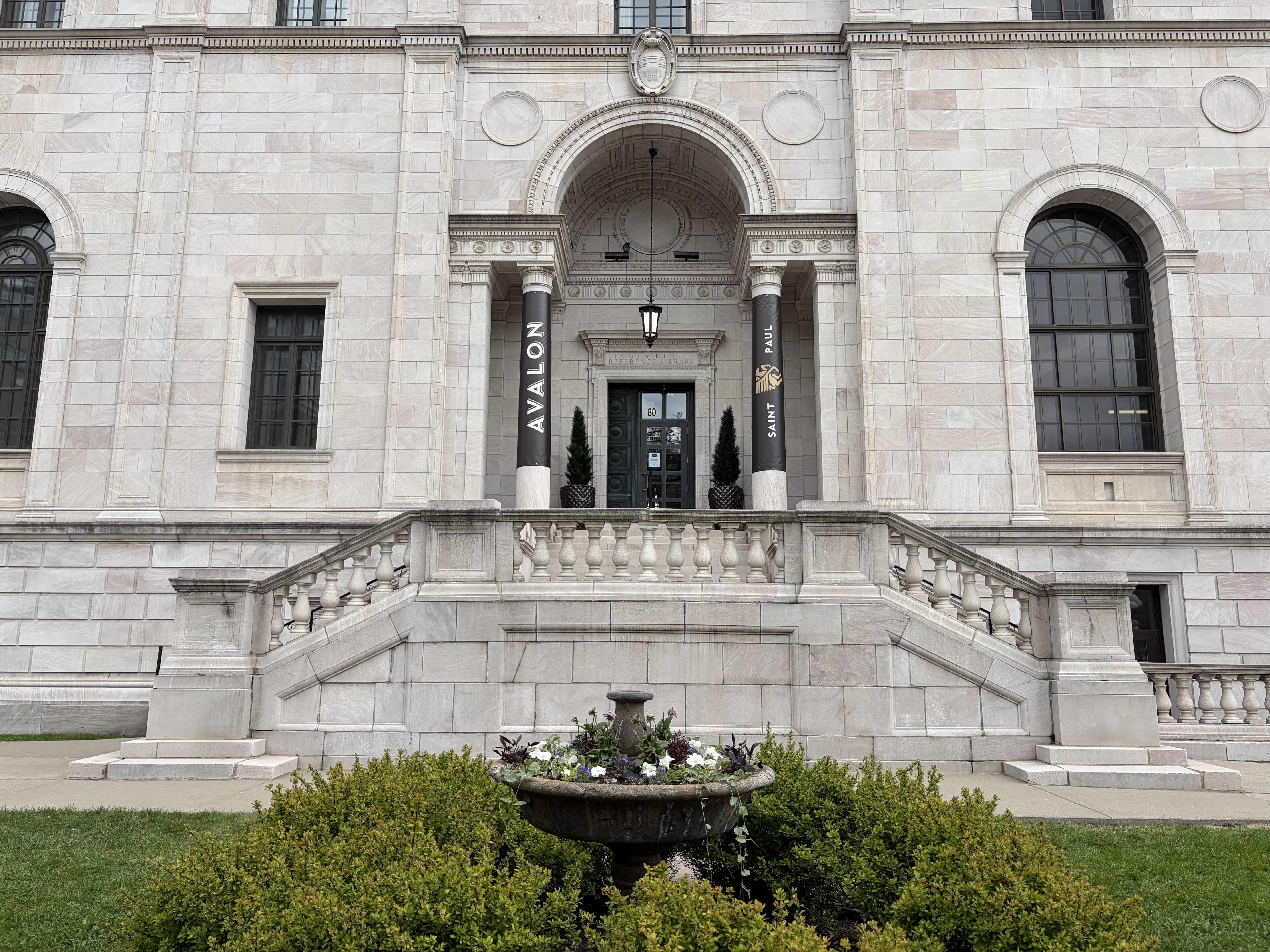 Front view of a grand beige marble building with a large central arched entrance. Two black banners read "AVALON" and "SAINT PAUL" beside the doorway, flanked by columns; a planter with flowers in front.