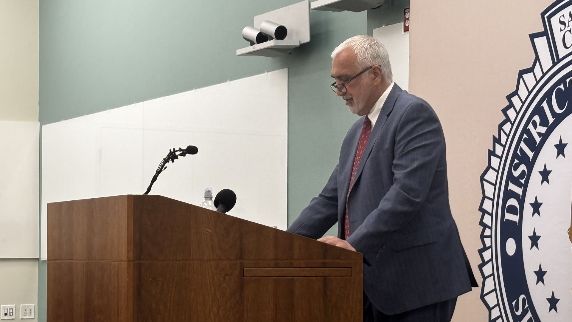 Man with white hair and glasses in a blue suit and red tie speaking at a wooden podium with microphones, beside a large emblem with stars and text on a beige wall.