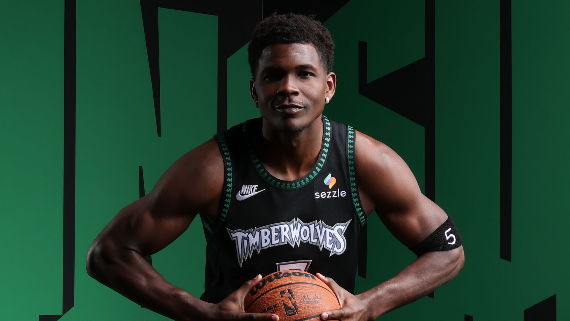 Anthony Edwards in black Timberwolves jersey with green trim, holding an NBA basketball against a green and black background.