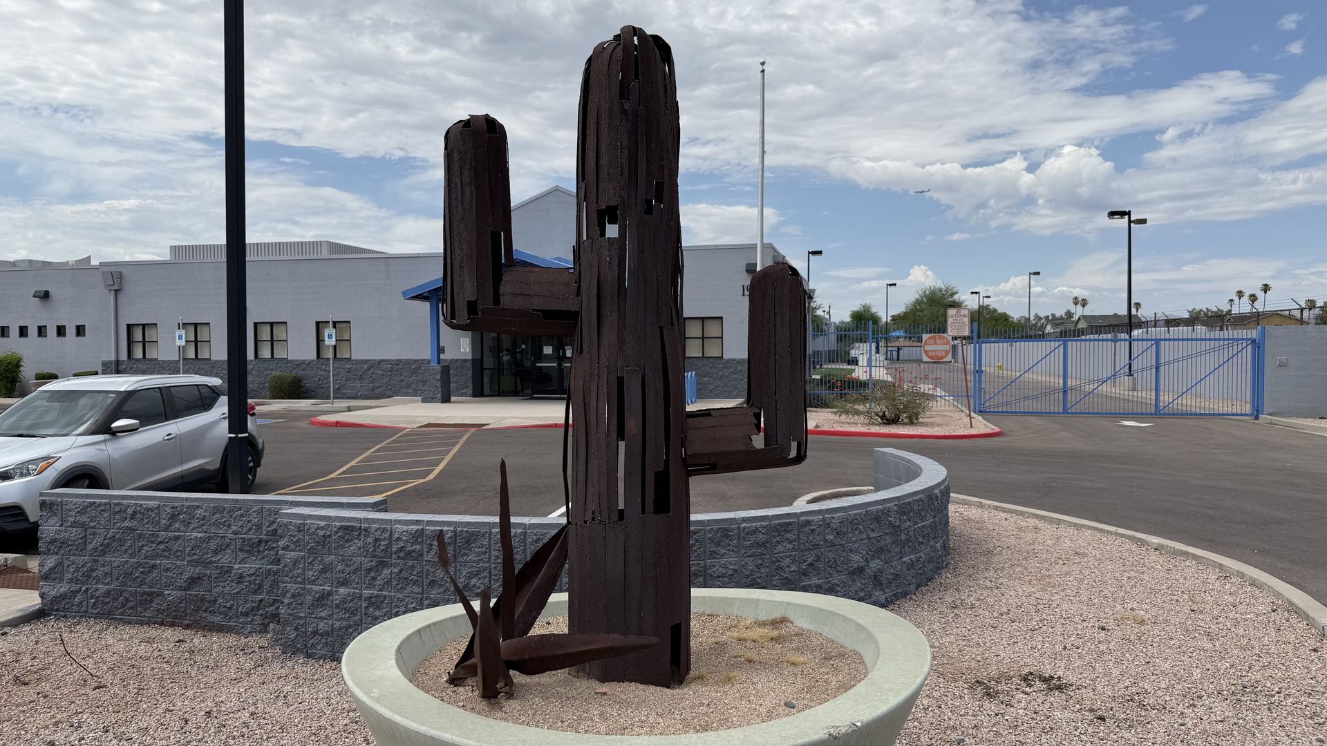 Rusty, metal sculpture shaped like a saguaro cactus rises from a circular concrete planter in front of a gray commercial building. A blue gate, parking lot, and a silver SUV are visible; a partly cloudy sky above.