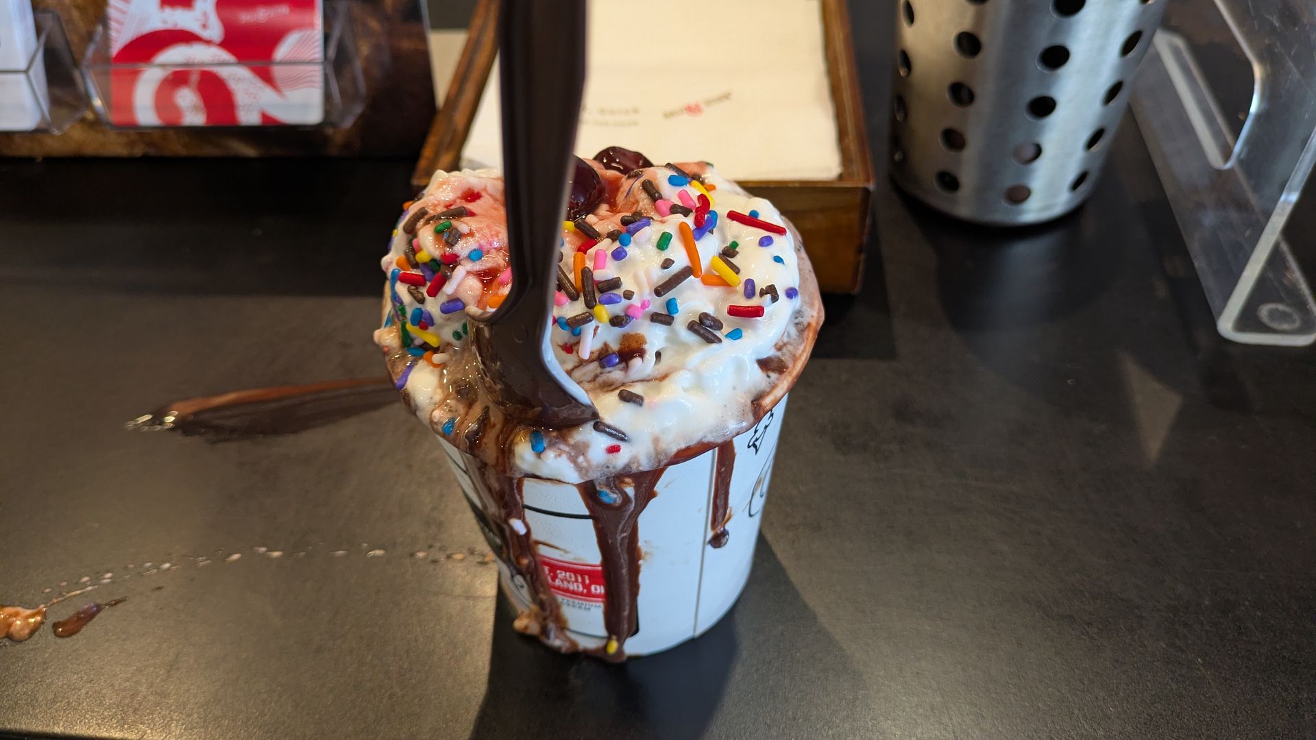 Milkshake in a white cup with white frosting, rainbow sprinkles, and chocolate drizzle; two dark chocolate sticks insert from the top, on a dark counter beside a metal utensil holder.