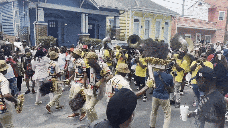 Street parade with musicians playing brass instruments, wearing colorful outfits and hats, surrounded by a crowd near blue and yellow houses on a sunny day.