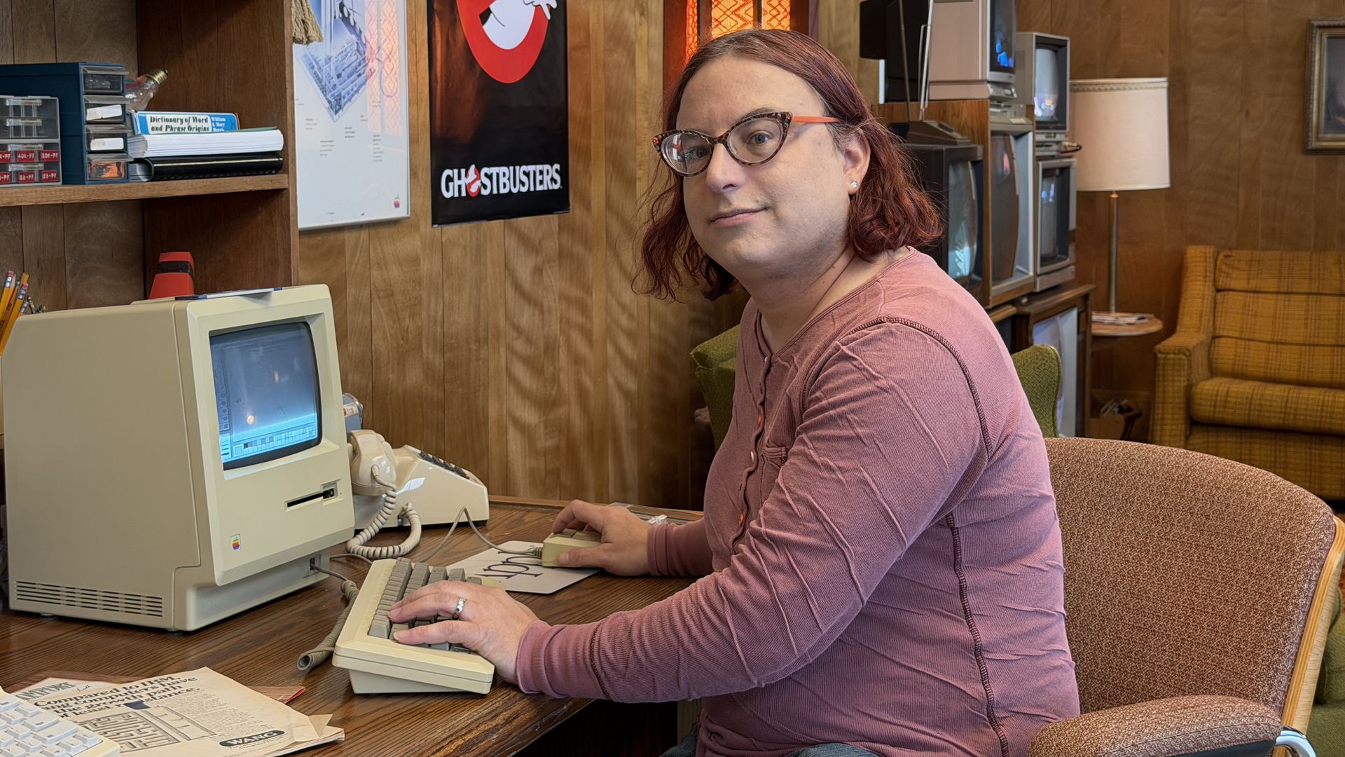Portrait of Axios' Ina Fried sitting in front of an original Mac computer on a wood-paneled desk. Behind her are retro shelves, a "Ghostbusters" poster, and a tan plaid couch in a retro office.F