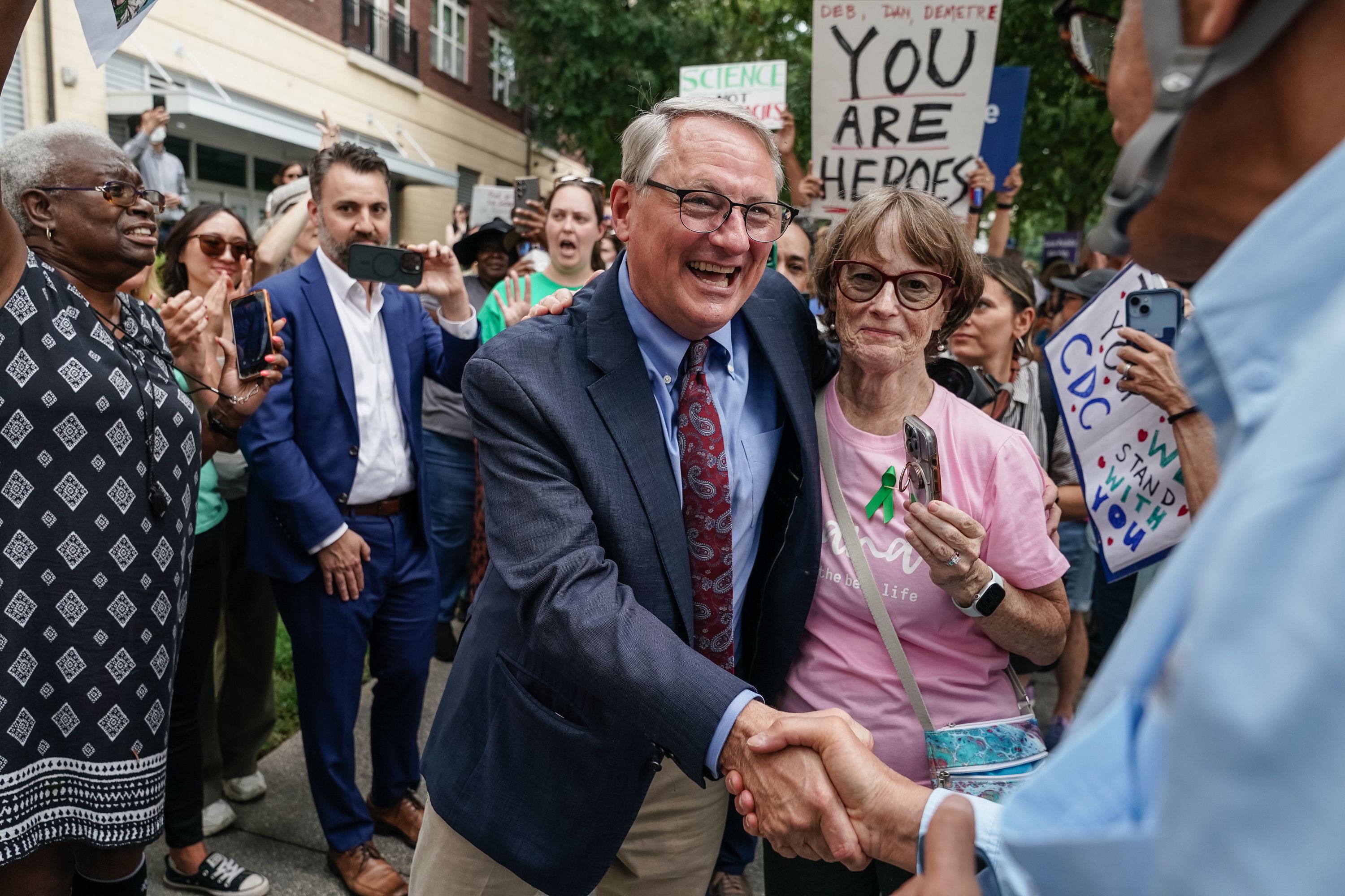 A man in a suit and glasses smiles while shaking hands with another person; a woman in a pink shirt holds a phone, surrounded by a crowd holding signs like "YOU ARE HEROES" at an outdoor event.