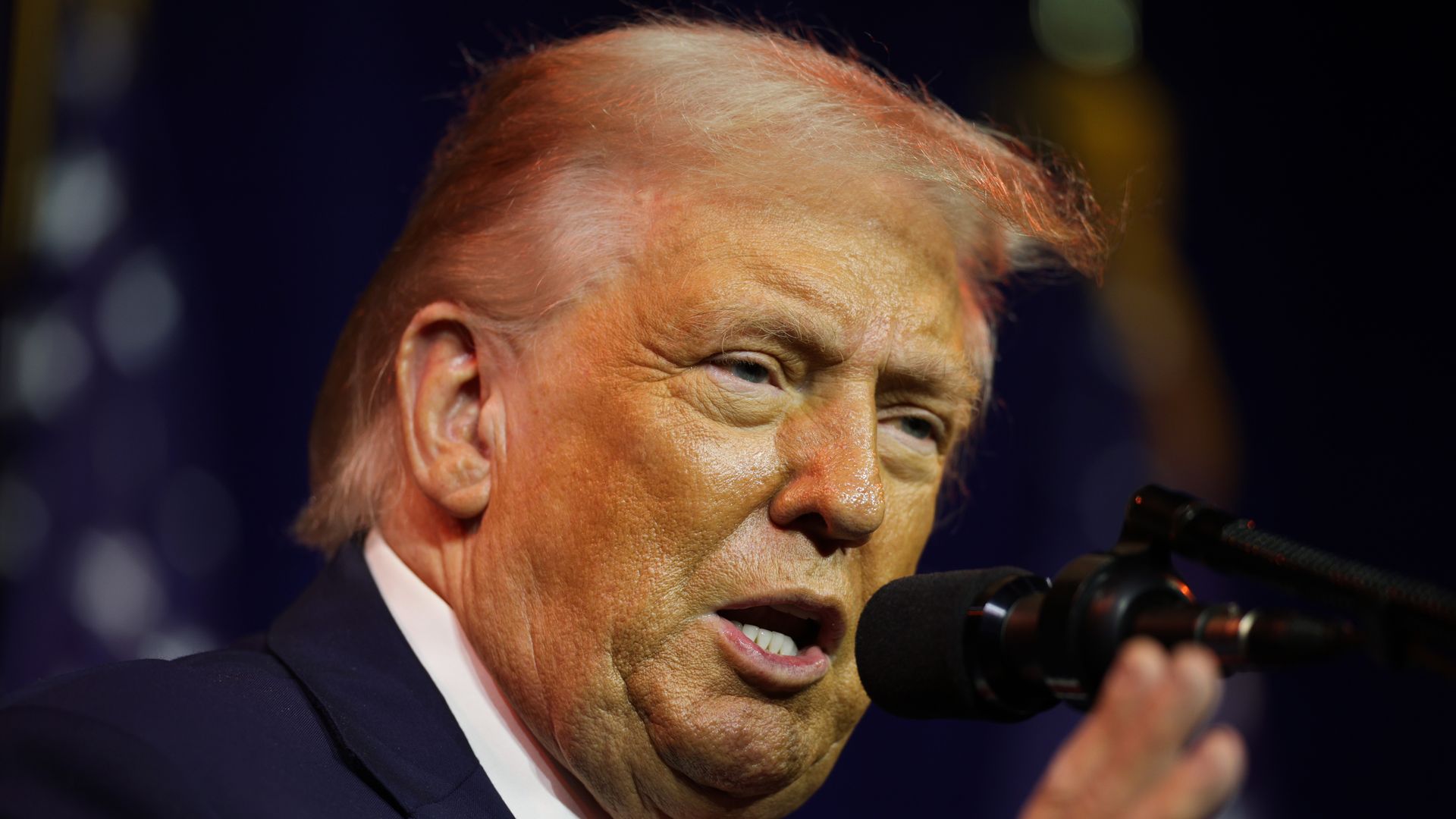 Trump speaking into a microphone with one hand raised, wearing a navy suit jacket, white collared shirt and red tie