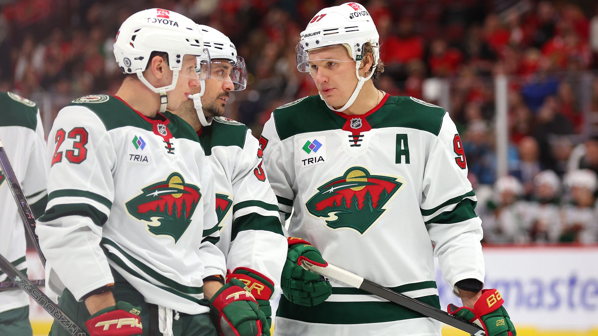 Three hockey players wearing white jerseys and helmets with green and red logos talk to each other on the ice during a game