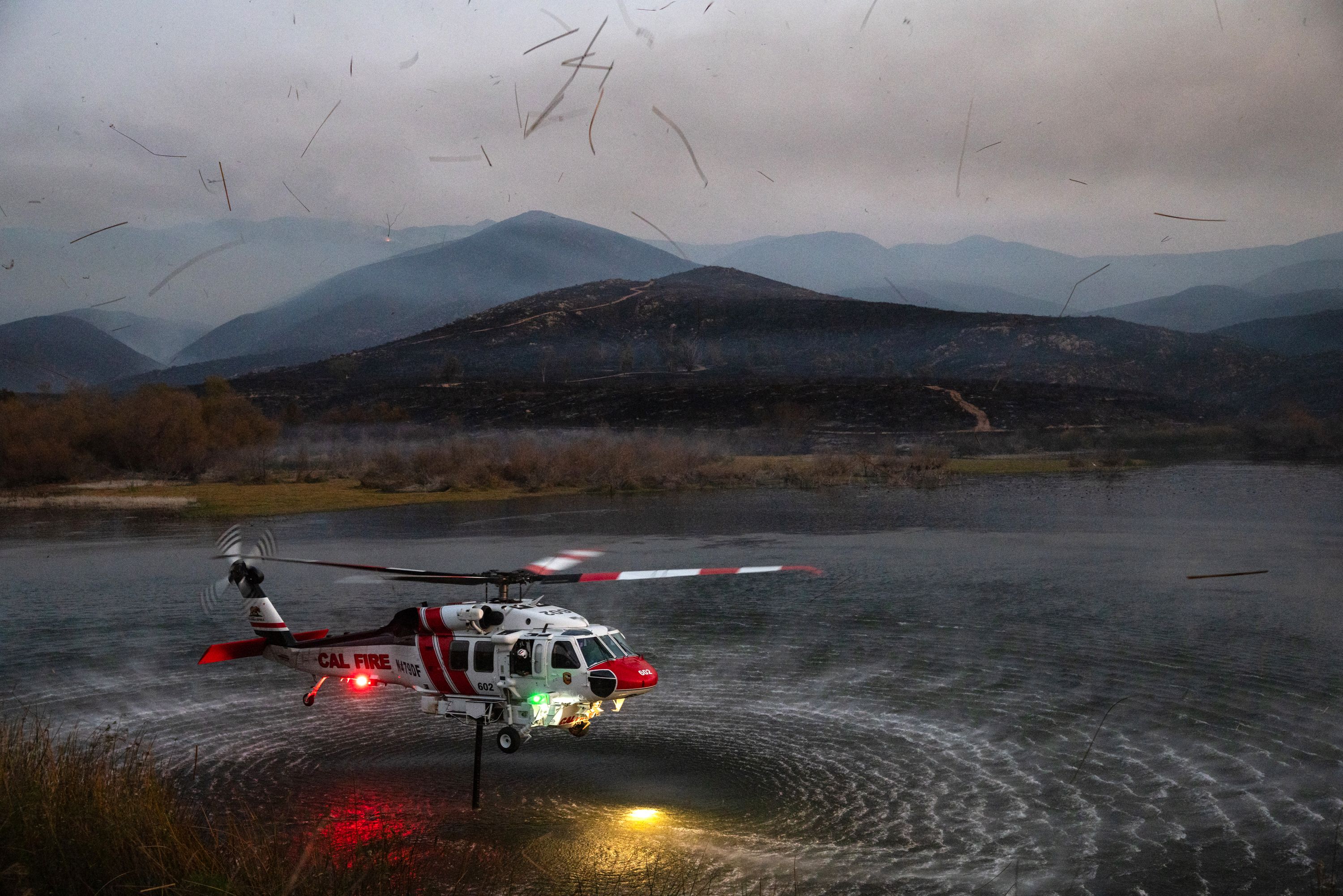 Overcast mountain range with layered blue ridges; in the foreground a CAL FIRE helicopter on a wet landing near the shore, red and white body with spinning rotor, water reflecting lights.