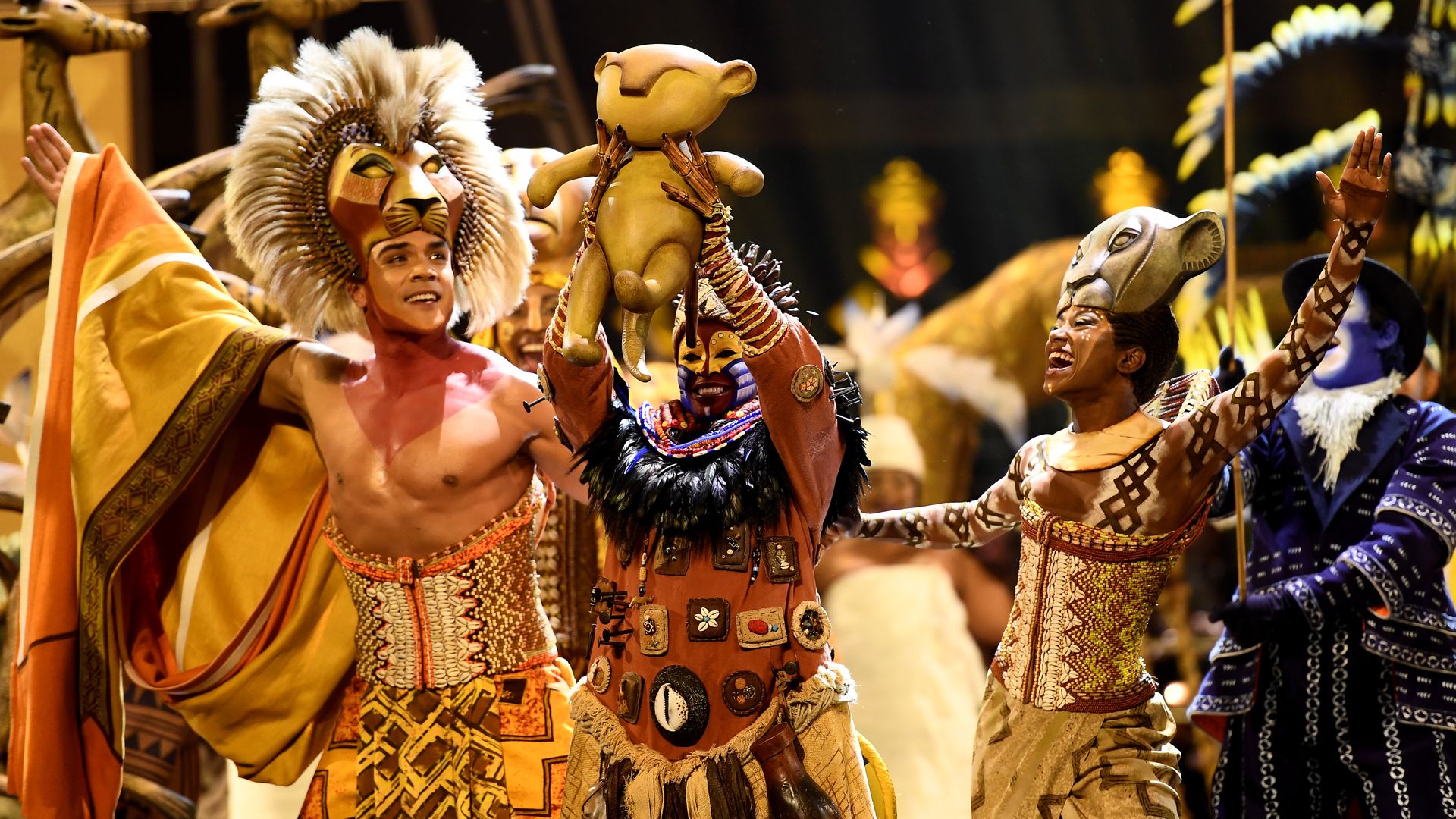 Image show performers in costume for "The Lion King." They are holding a baby Simba in the air.