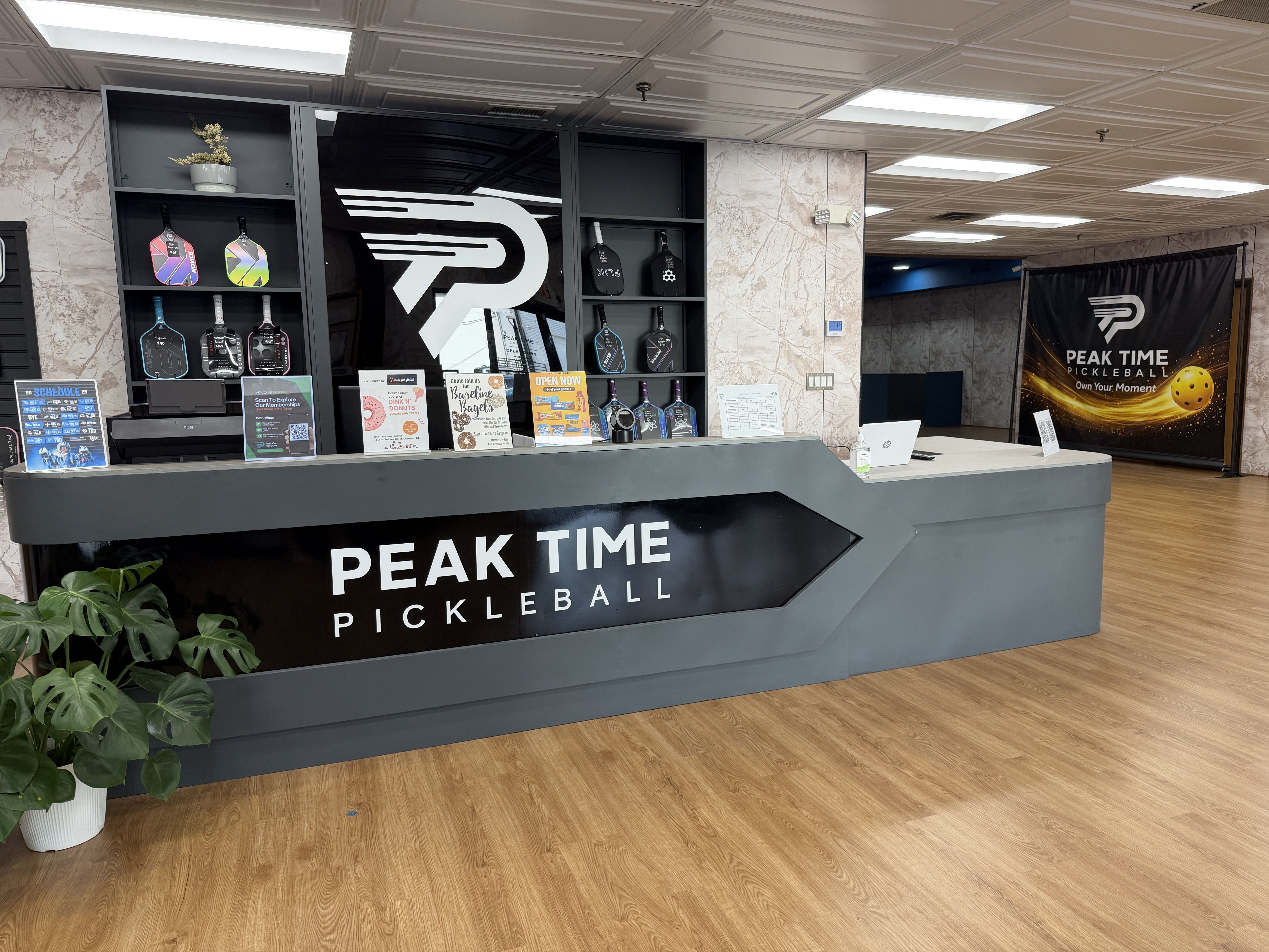 Interior of a Peak Time Pickleball shop with a gray counter, shelves of colorful paddles, a large white logo, marble walls, a potted plant, and a black banner reading Peak Time Pickleball.