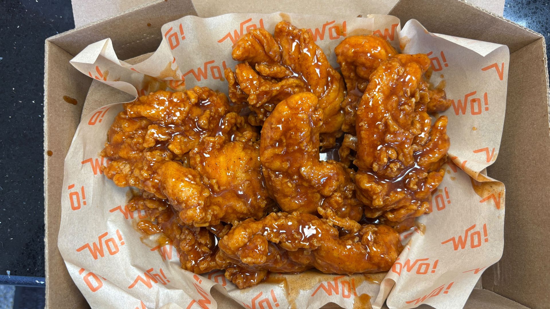 Seven pieces of crispy fried chicken wings coated in a shiny, orange-brown sauce, served on white paper with orange "WO!" text inside a brown cardboard takeout box.