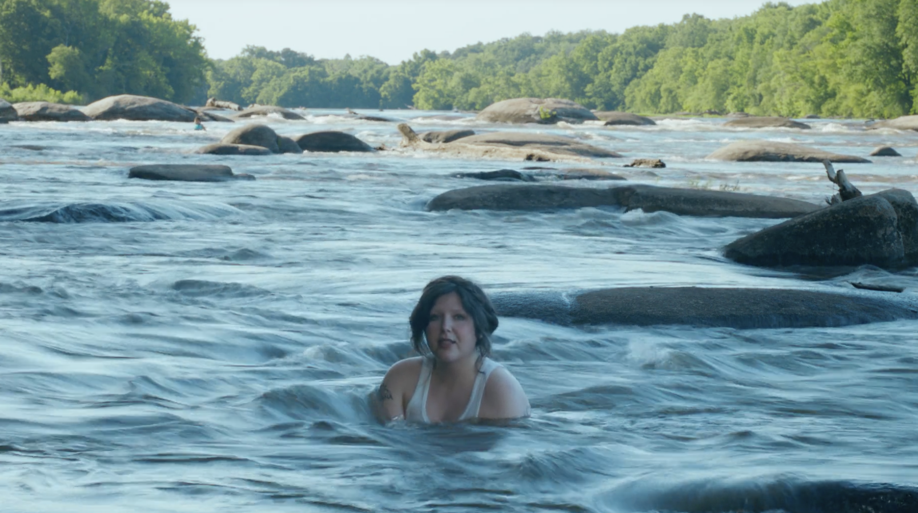 A woman in a white tank top stands waist-deep in a fast-flowing river, surrounded by large smooth rocks, with a lush green forest along the distant riverbank.