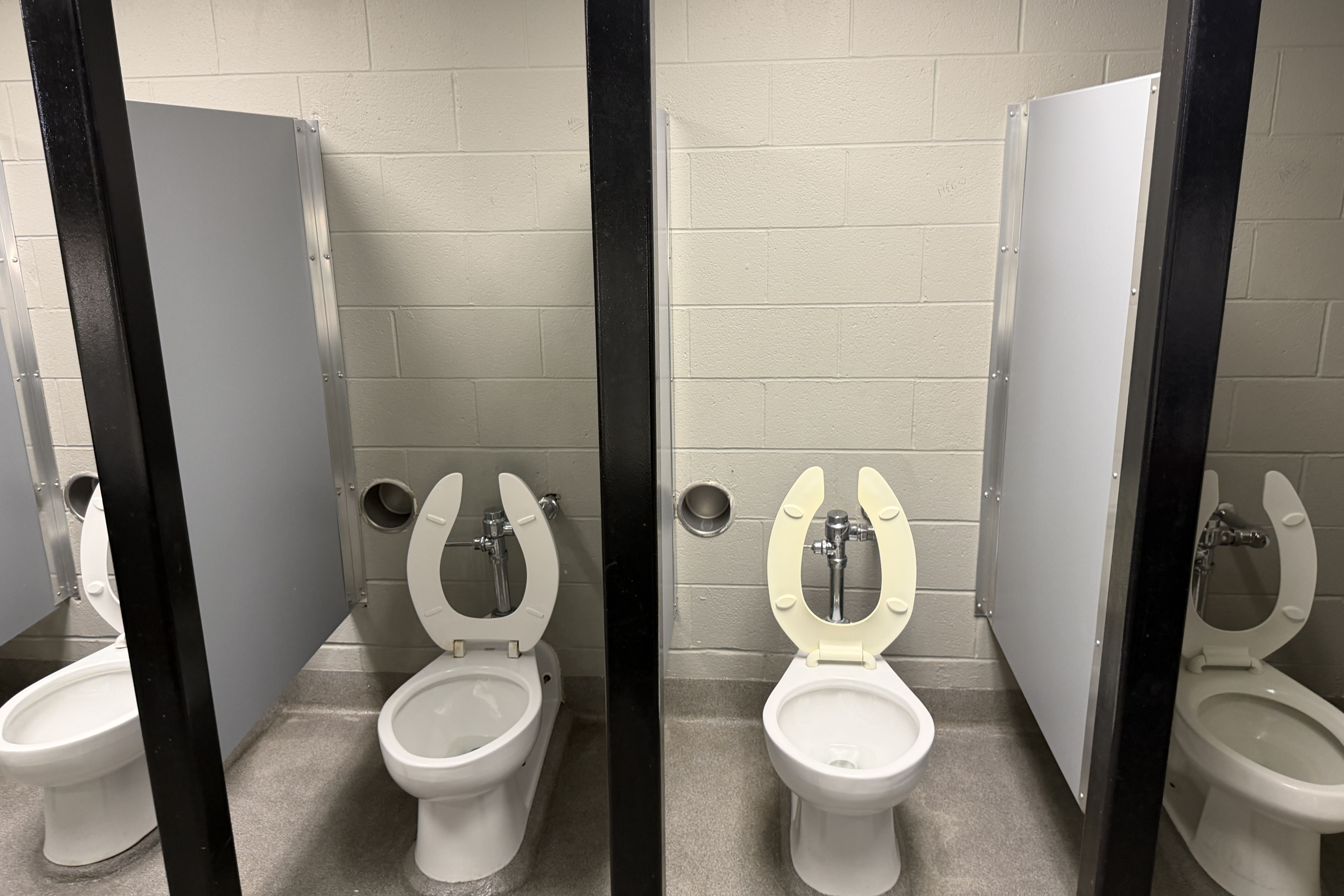 Public restroom with three stalls: white toilets with seats up, gray doors, and black metal divider frames; beige cinder-block walls and metal panel partitions.