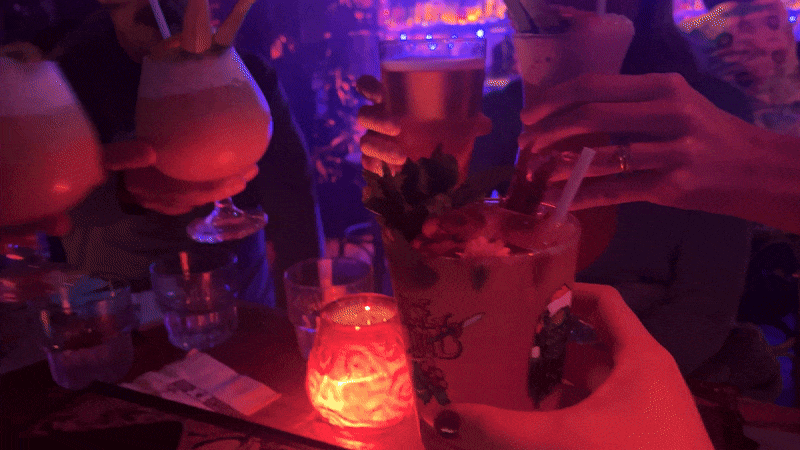 A video shows four people cheers-ing tiki-themed cocktails at a dark bar. 