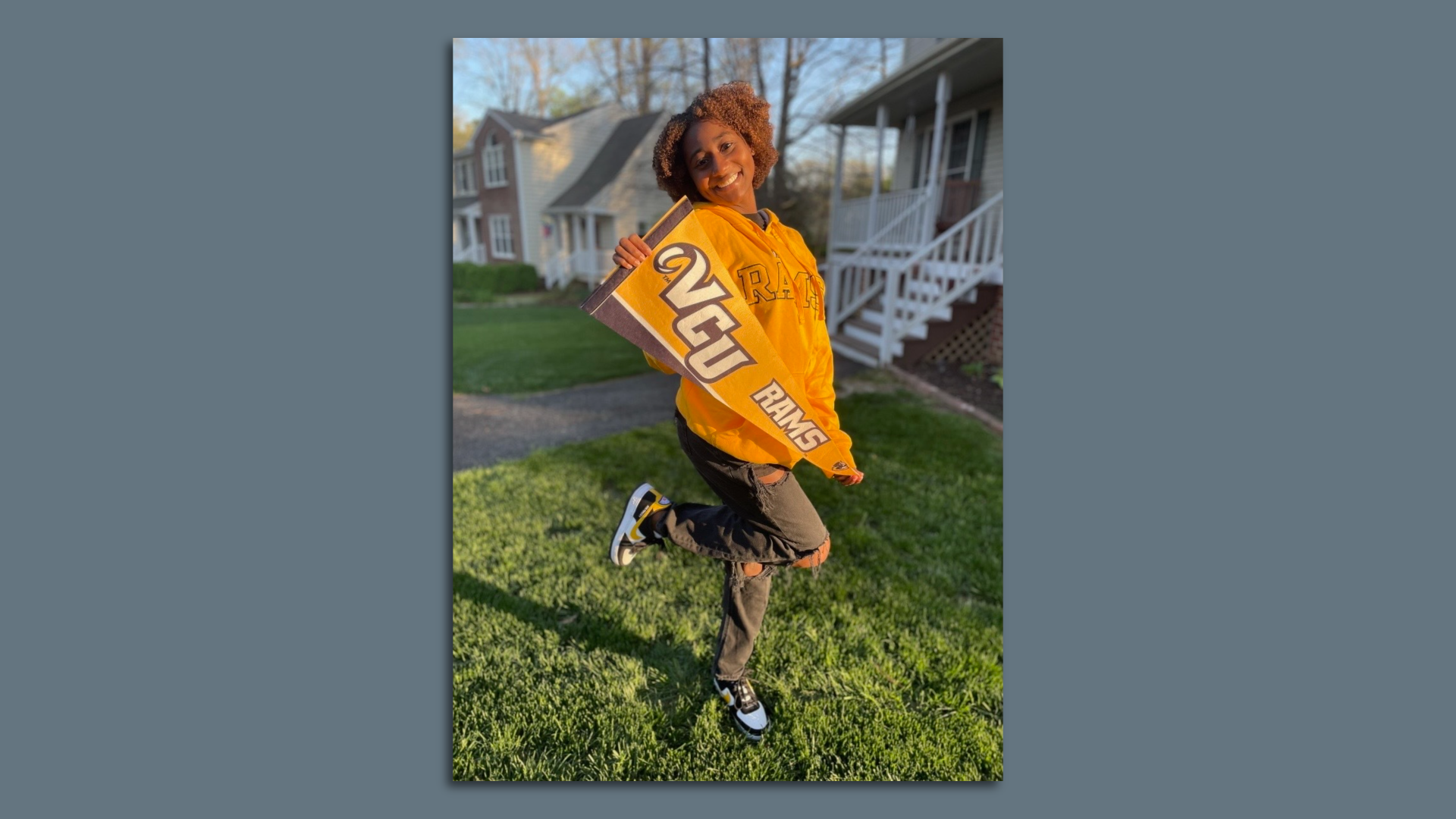 A photo of a woman with a yellow VCU sweatshirt holding a VCU Rams flag posing in front of a house
