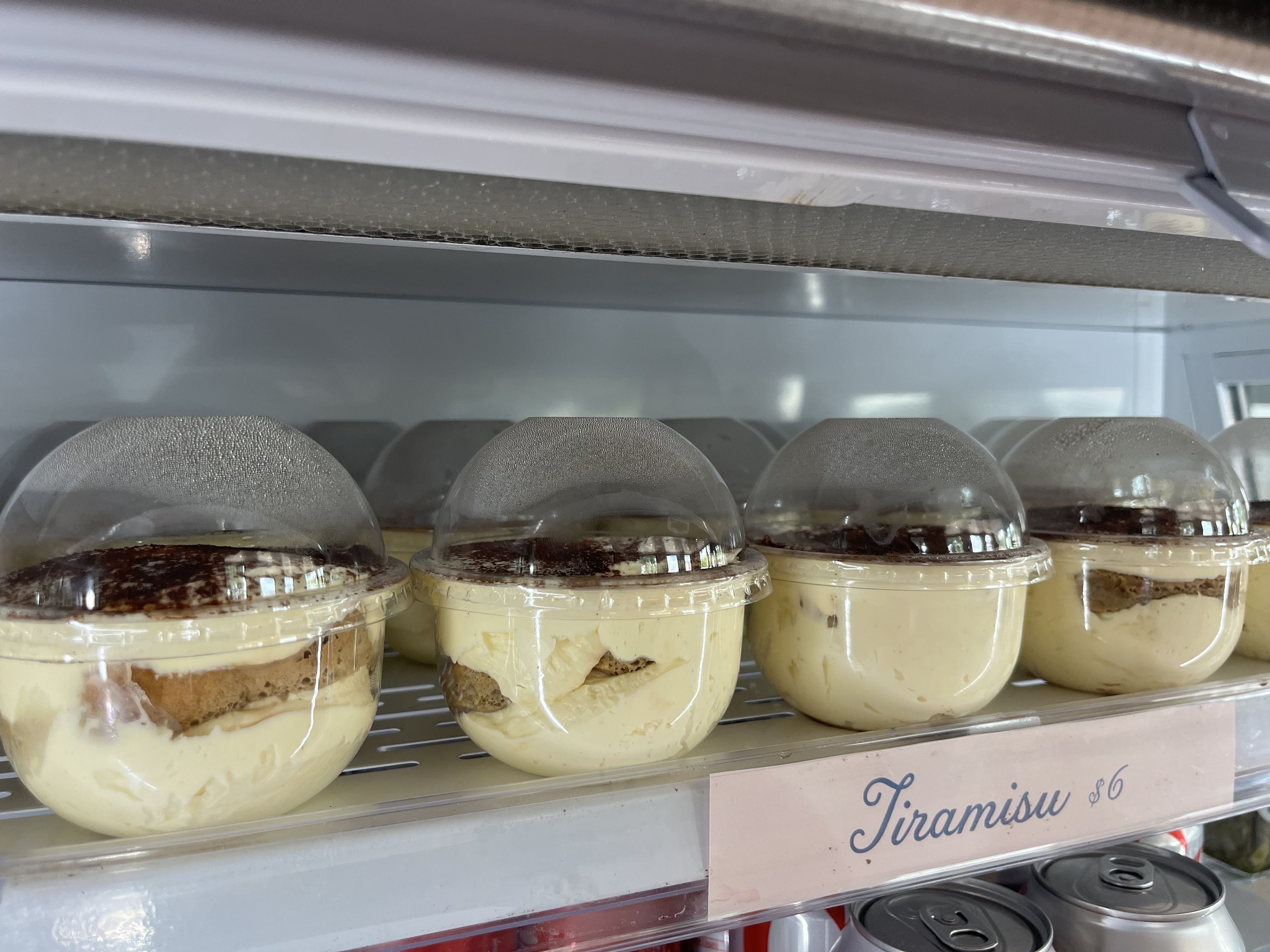 Plastic cups of tiramisu dessert with creamy layers and cocoa powder, displayed in a refrigerated shelf with a price tag of $6.