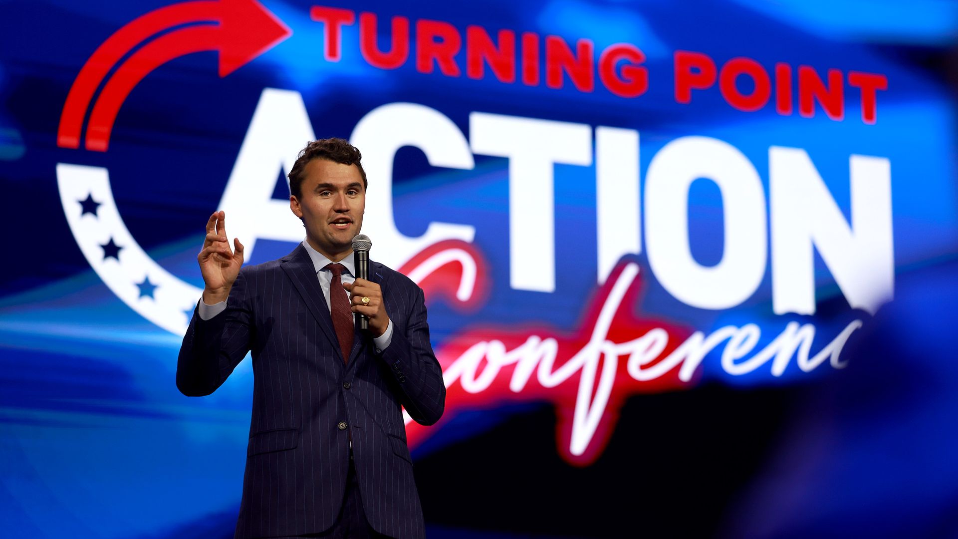 Charlie Kirk, wearing a blue suit and holding a microphone, speaking in front of a large blue, white and red Turning Point Action Conference backdrop.