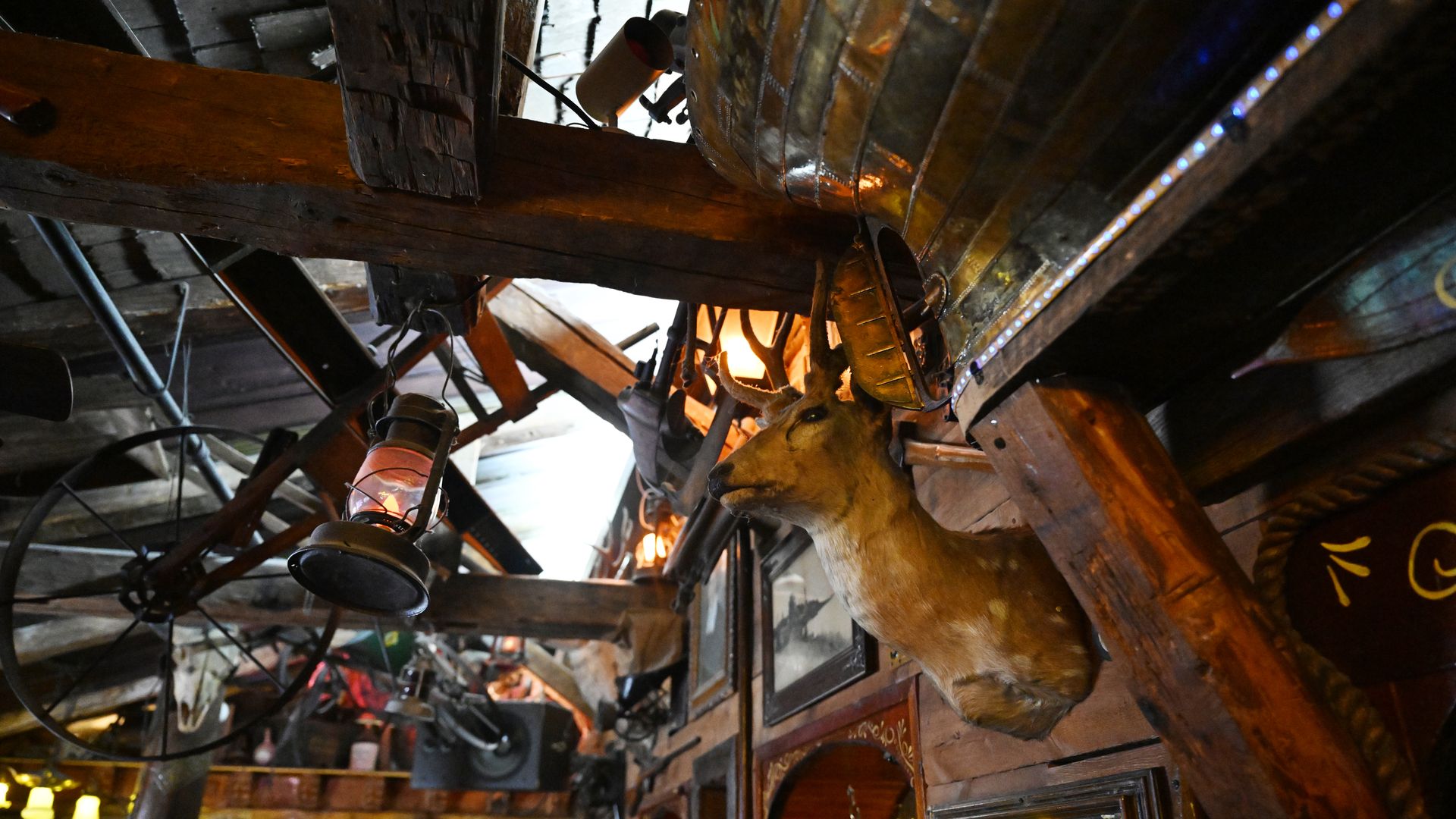 Rustic tavern interior with dark wooden beams, a mounted deer head near the ceiling, glowing lanterns, string lights, and vintage framed mirrors on weathered walls.