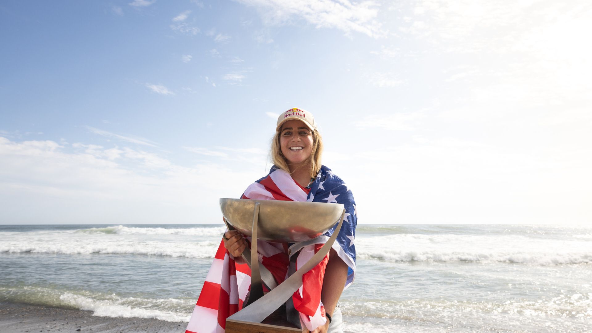 A woman draped in an American flag stands at the beach holding a big bowl/trophy.