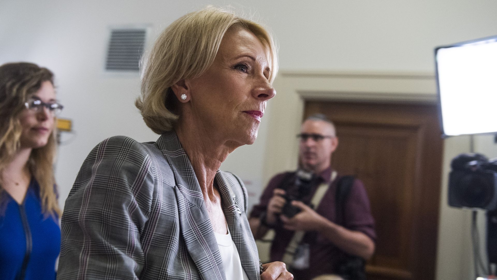 In this image, DeVos walks down a hallway with cameras and lights nearby.