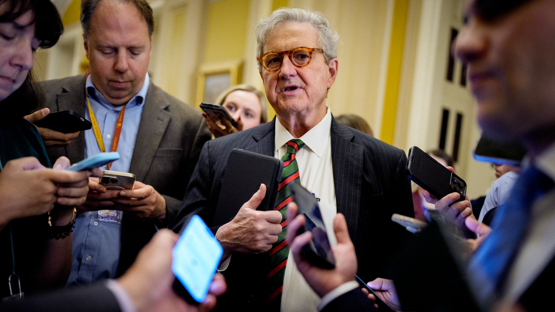 Sen. John Kennedy clutches a tablet in one hand as he's surrounded by reporters holding out cell phones while he speaks.