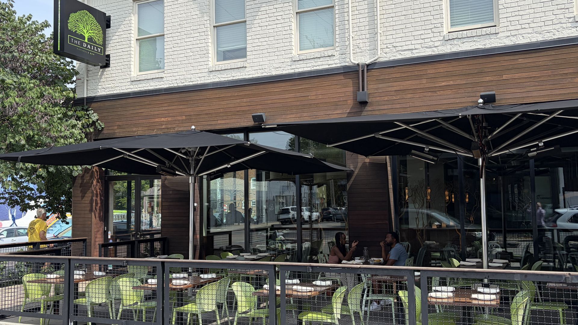 Exterior of a cafe called The Daily with a bright green tree logo on a black sign mounted to a white brick wall; outdoor seating with lime-green chairs beneath large black umbrellas.