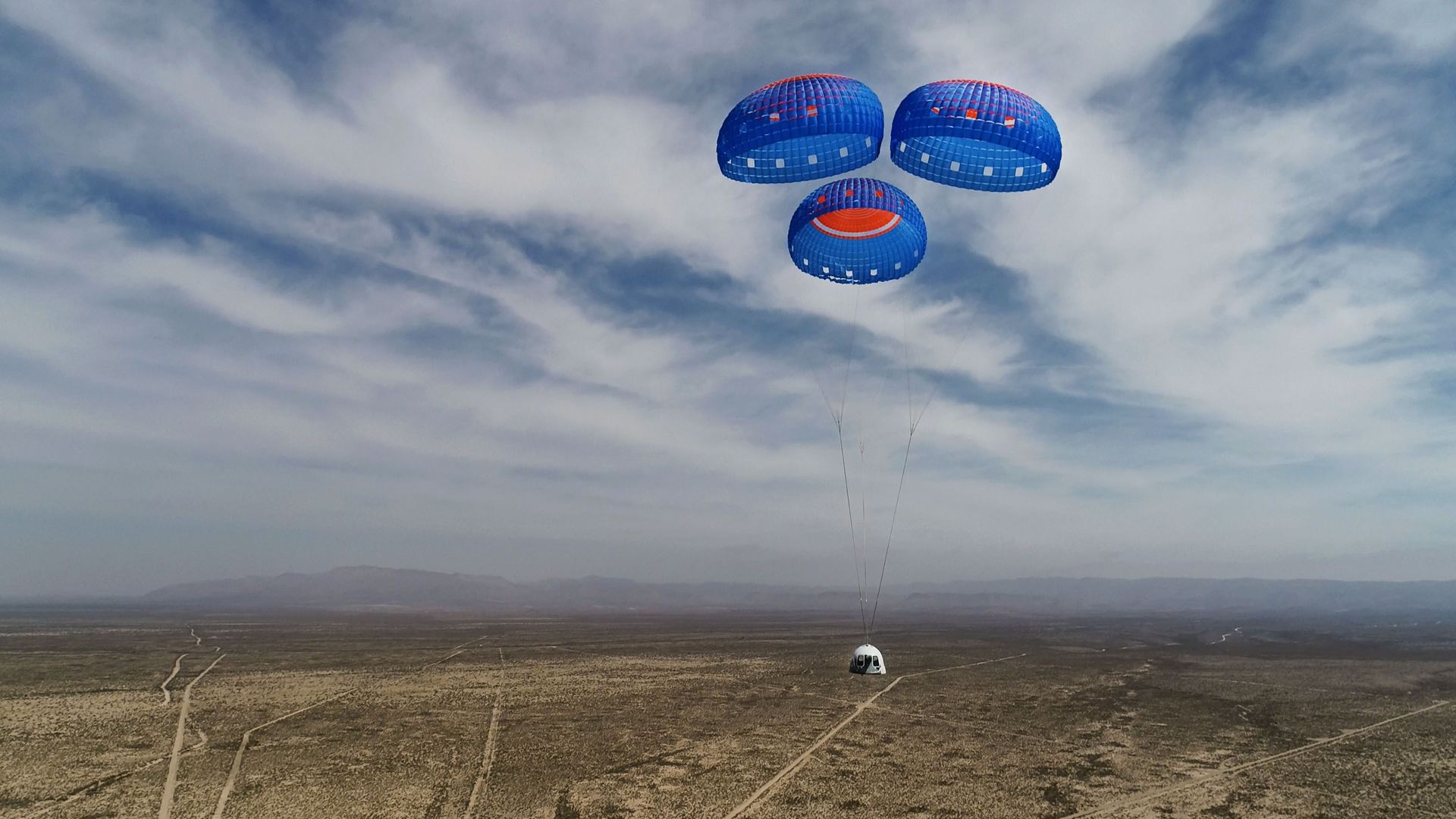 A Blue Origin capsule descending back to Earth against blue skies and clouds under three blue and red parachutes