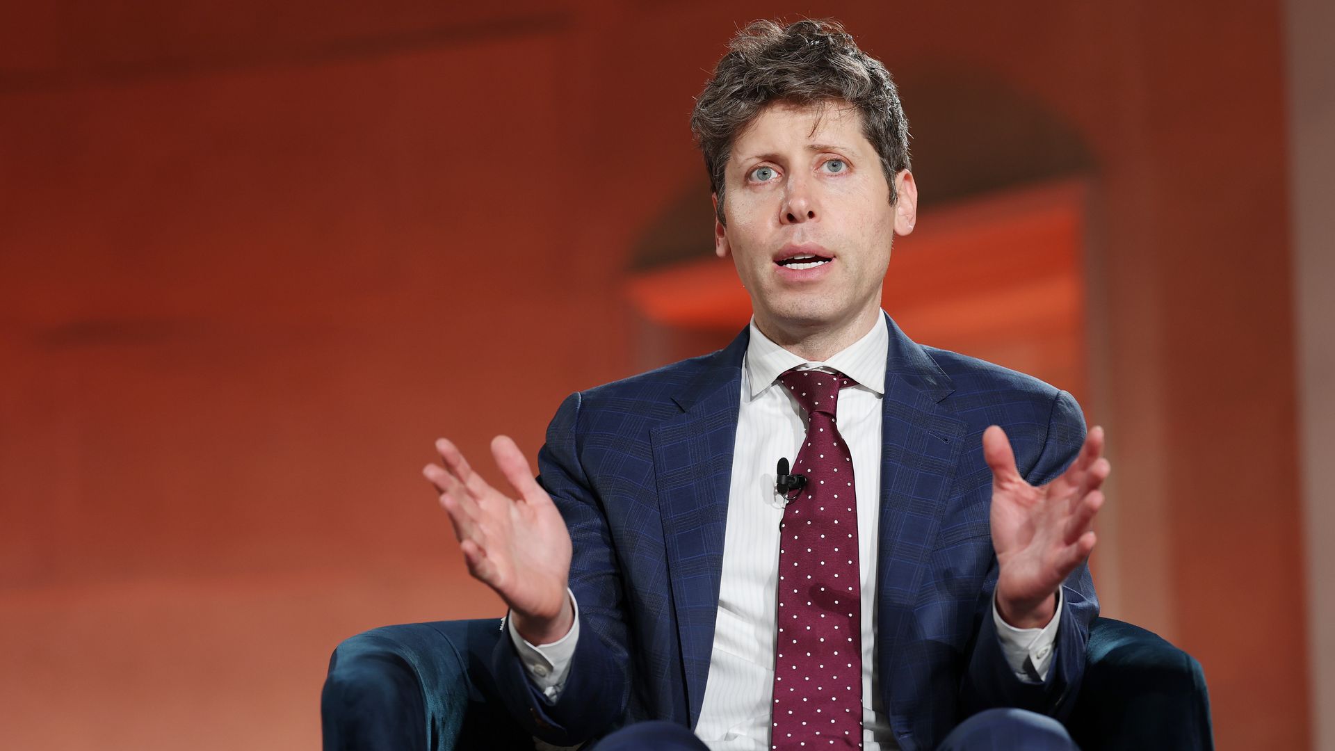 A man in a suit sitting on a chair on stage holding his hands out while talking