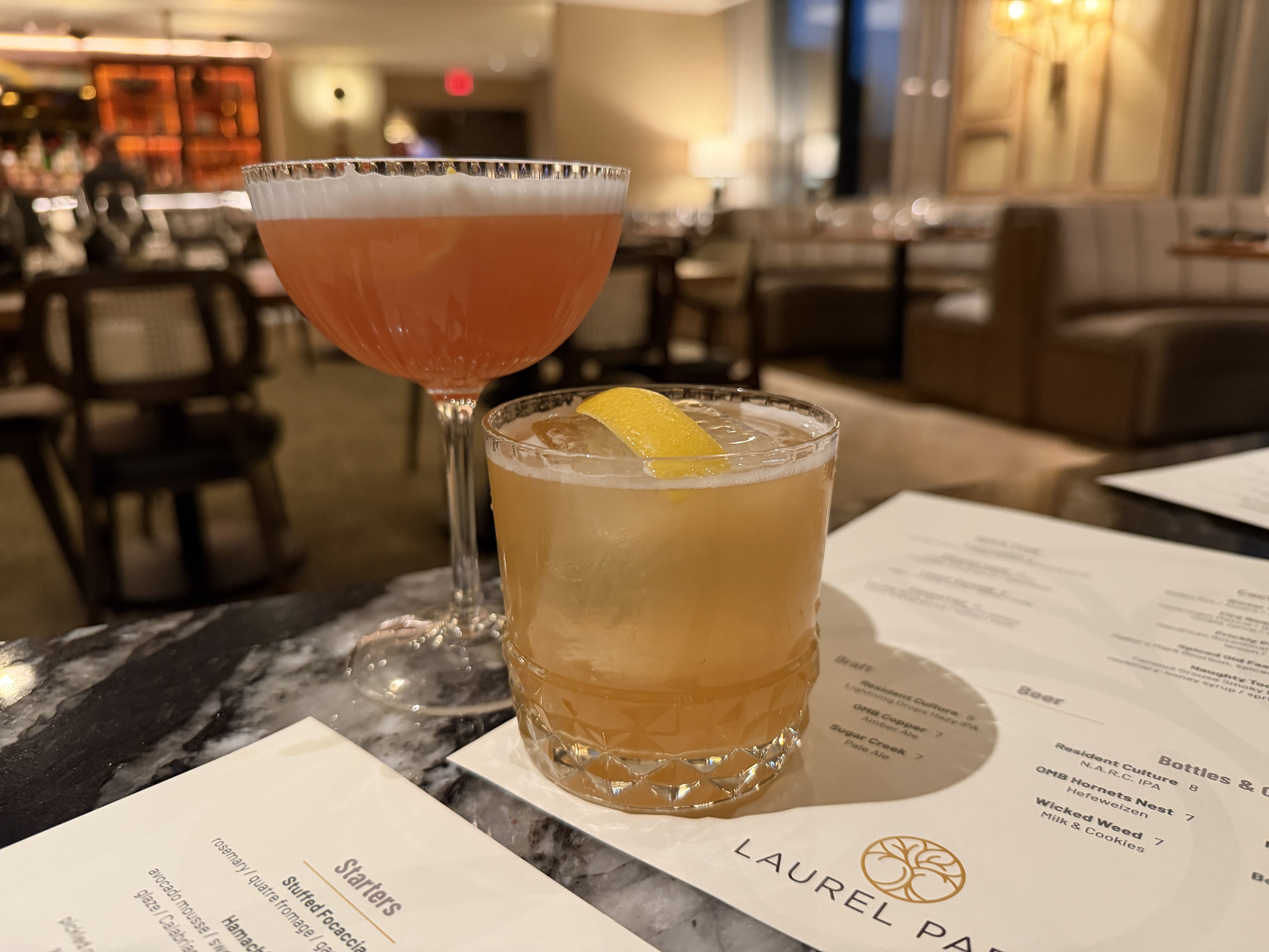 Two cocktails on a marble table in a cozy, softly lit restaurant; one pink drink in a coupe glass and one amber drink with a lemon twist in a short glass, with menus from Laurel Park.