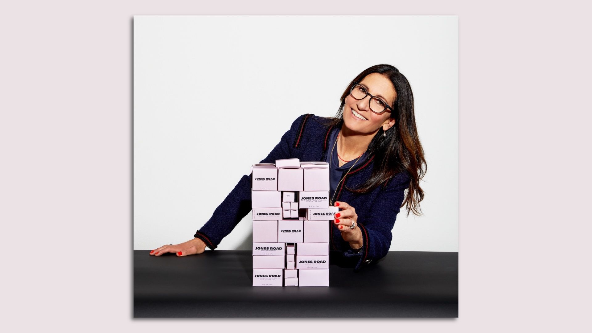 Woman with long brown hair and glasses sitting behind a stack of light pink boxes.