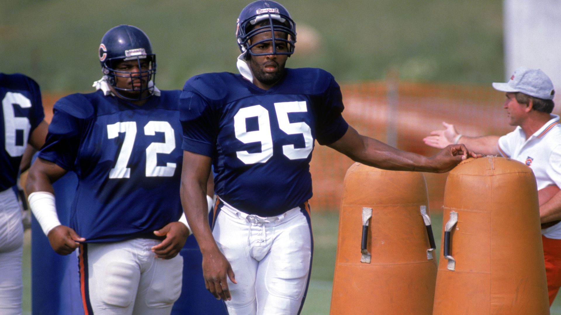 Three football players in blue jerseys sprint past orange practice dummies on a field; visible numbers include 95 and 72, as a coach in a white shirt and cap gestures to them.
