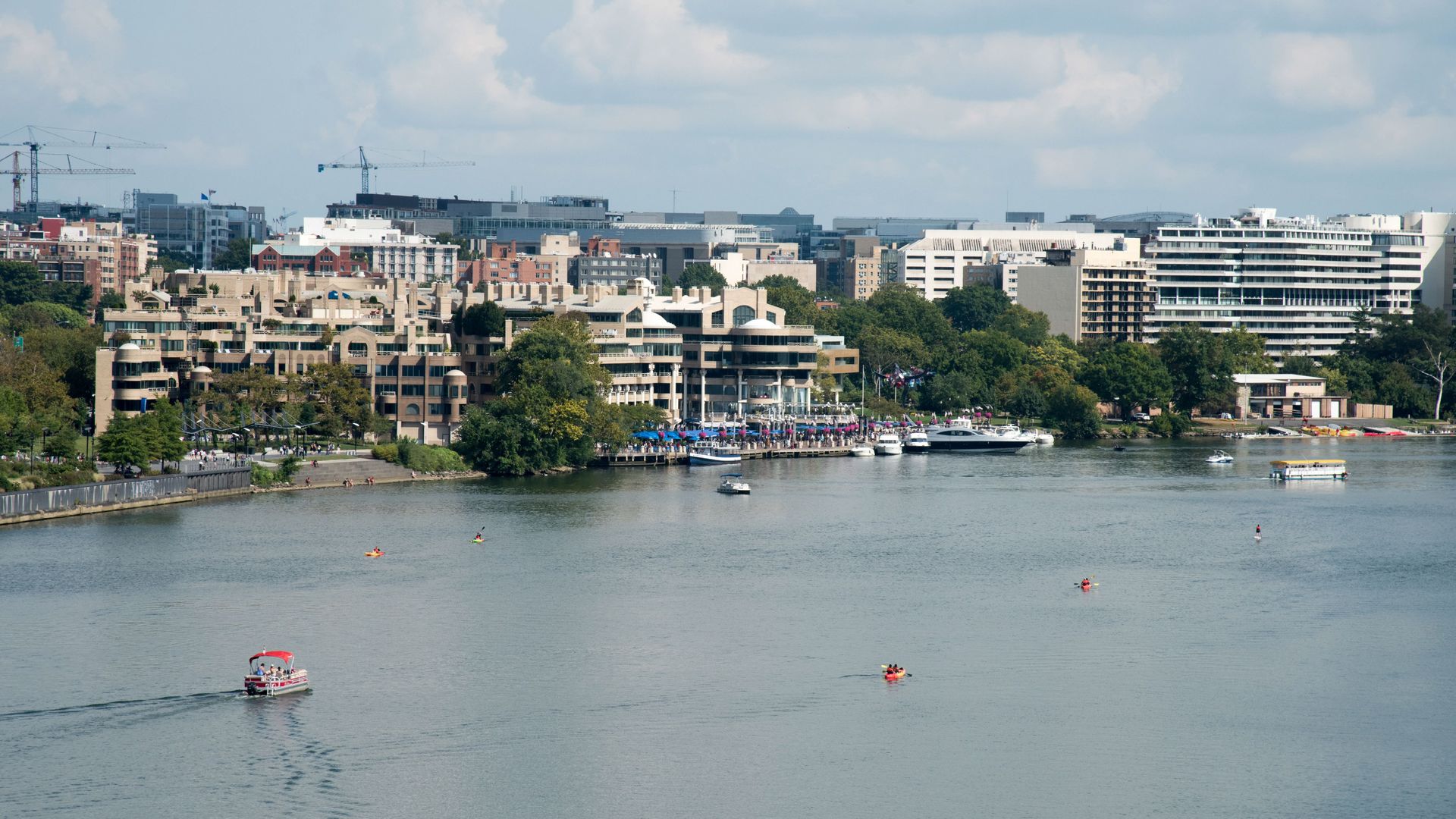 River with small boats and kayaks, shoreline with trees and crowds near a beige multi-story building, cityscape background under cloudy sky.