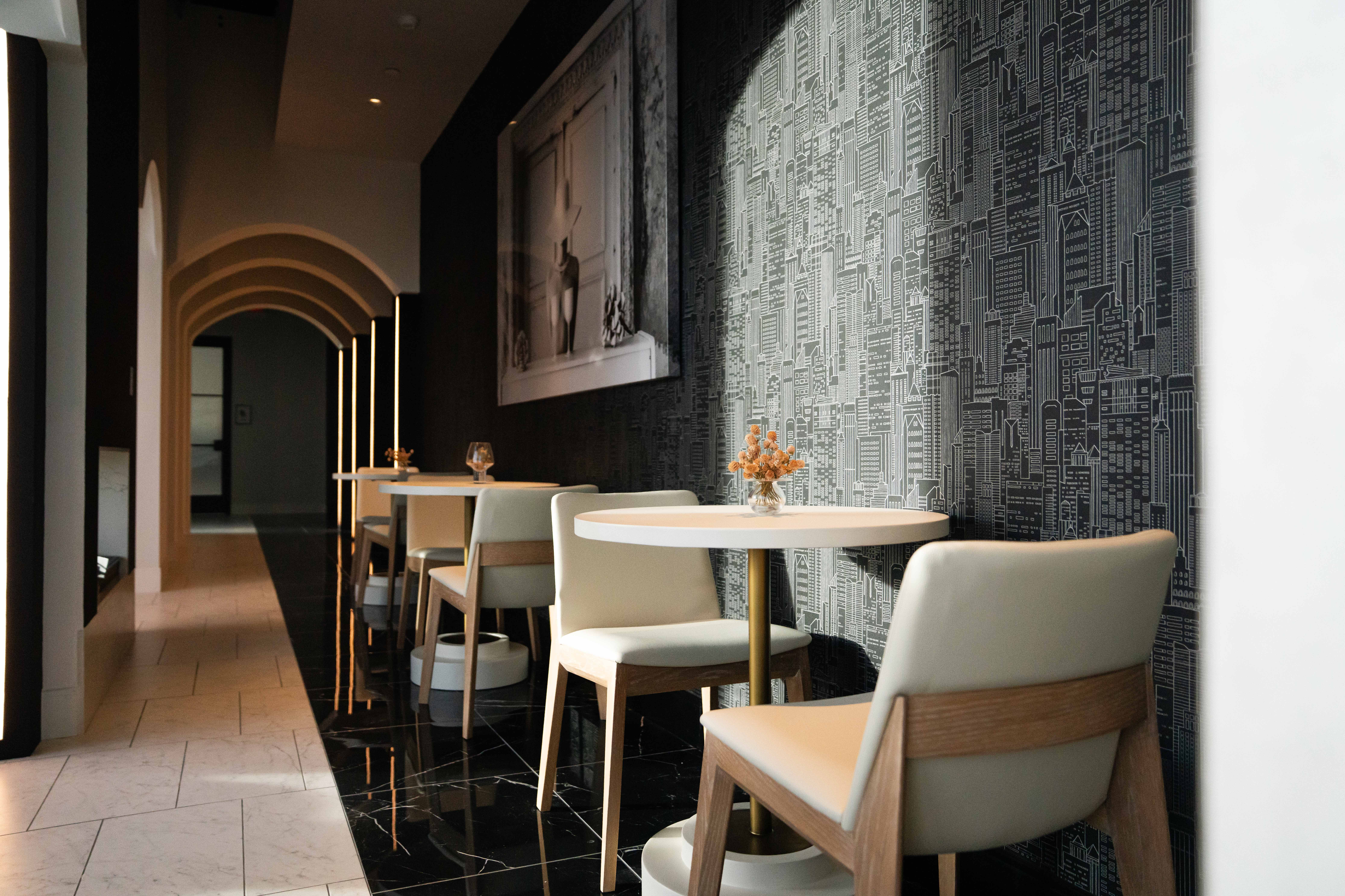 Modern cafe interior with round white tables, beige chairs, black cityscape wallpaper, arched ceiling lights, and a small flower vase on the foreground table.