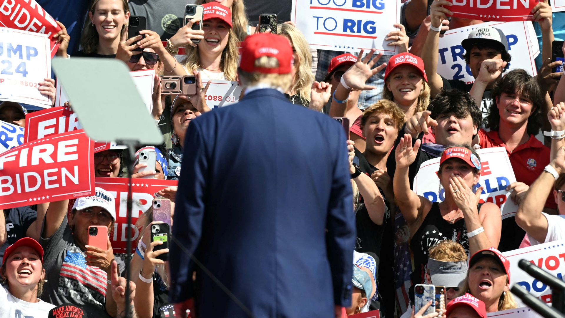 A man in a blue suit and red ball cap, with his back to the camera, faces a large crowd of cheering fans holding political signs, many of which have pro-Trump or anti-Biden messages. 