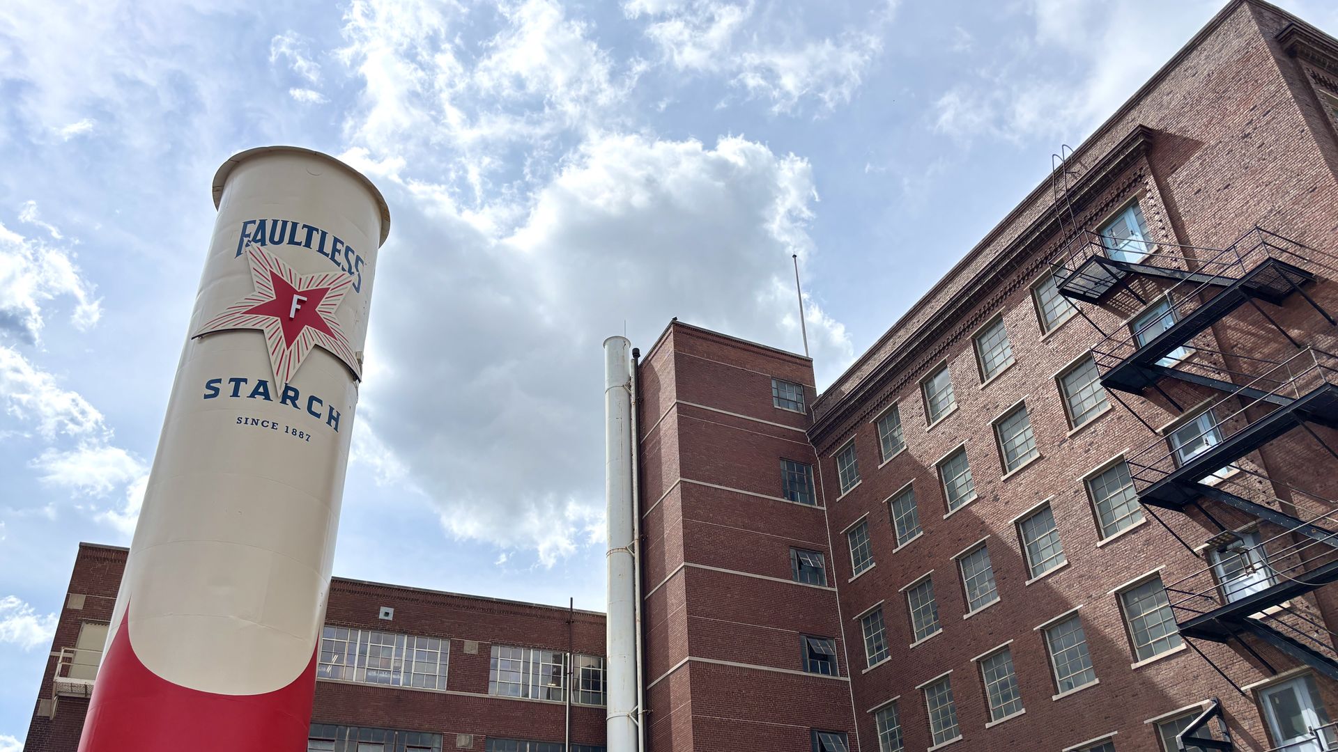 Tall white silo with the words "FAULTLESS STARCH" and a red star logo, standing beside a red brick building with a metal fire escape, under a blue sky with white clouds.
