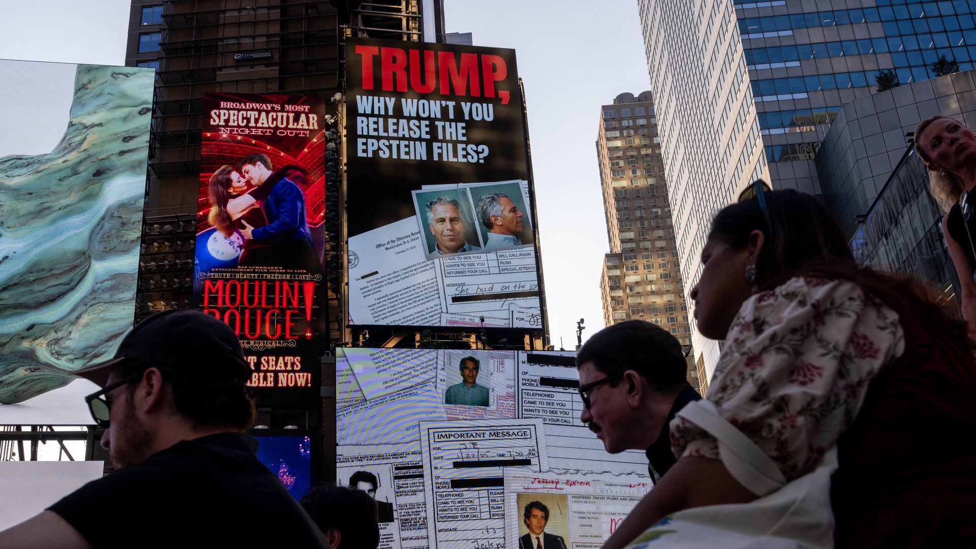 People in silhouette in a city square with large digital billboards, including one that says: "TRUMP, WHY WON'T YOU RELEASE THE EPSTEIN FILES?", with images of documents and photos.