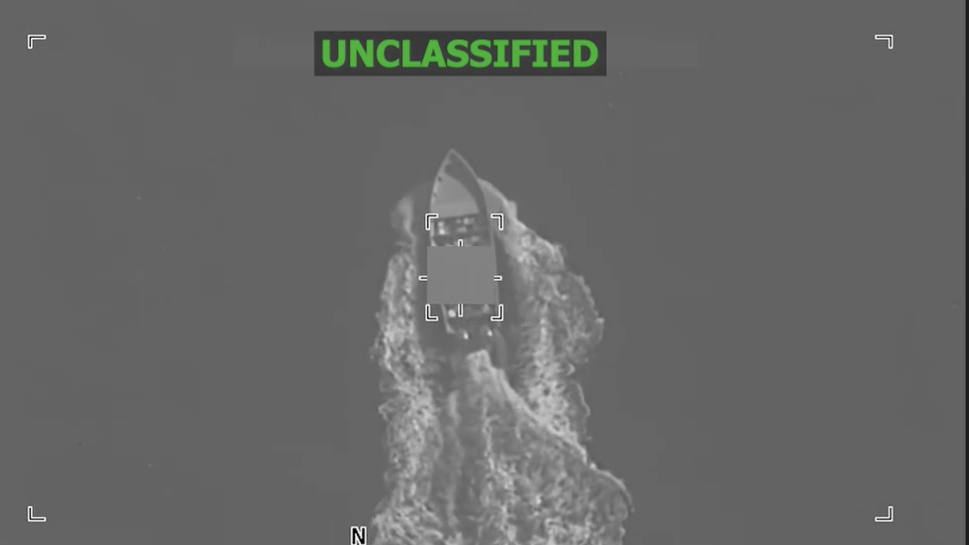 Black and white aerial view of a boat moving through water, creating a wake behind it. The image is marked "UNCLASSIFIED" in green at the top.