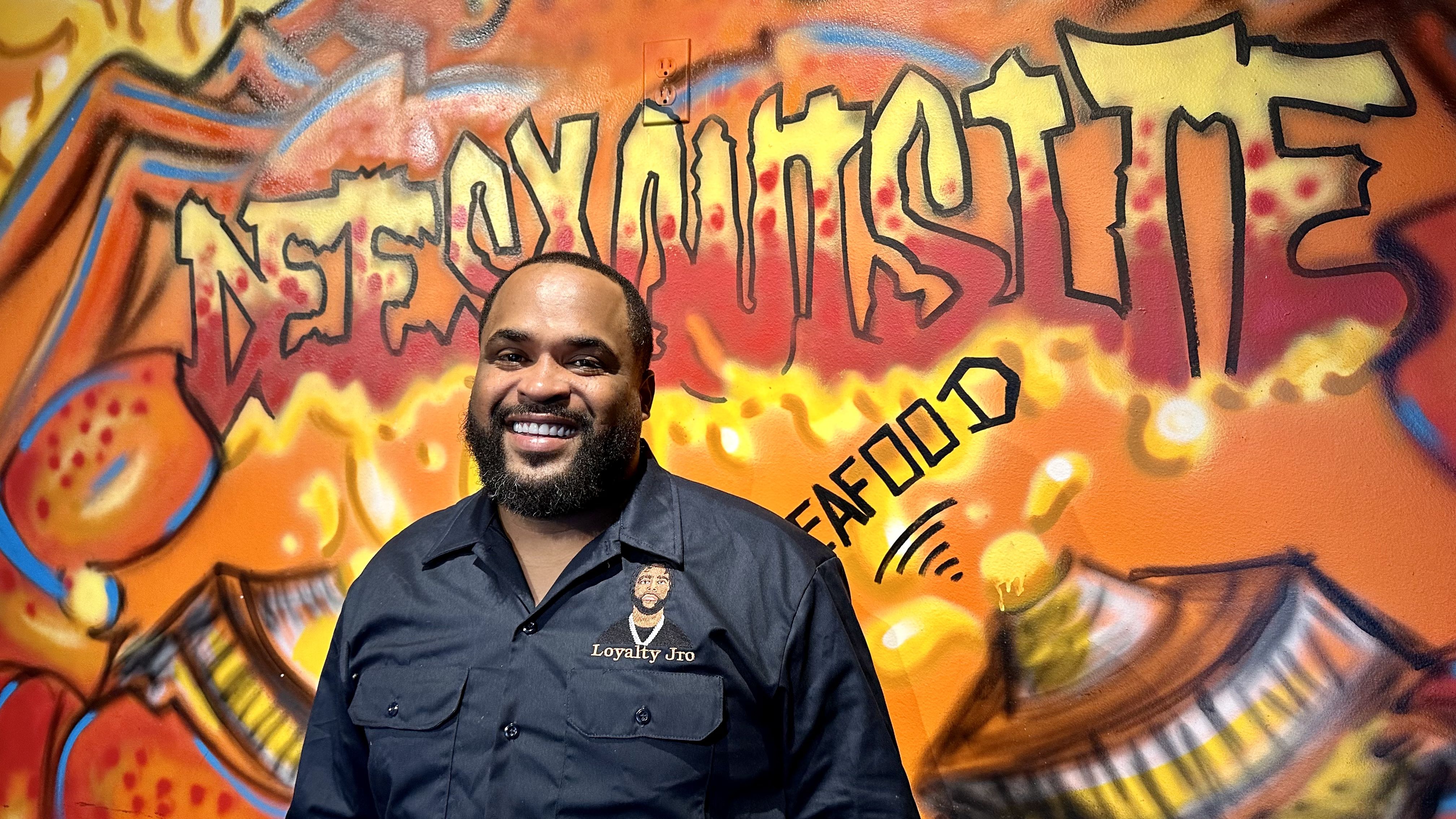 Photo shows Demond Matthews smiling in front of a mural in his restaurant.