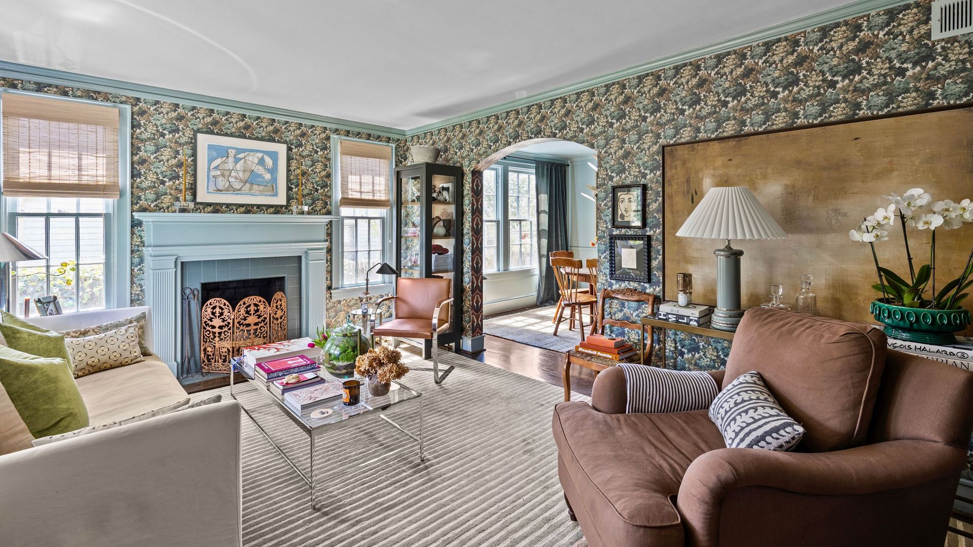 Cozy living room with floral wallpaper, light blue trim, beige sofa with green pillows, brown armchair, glass coffee table, and fireplace with blue mantel and decorative screen.