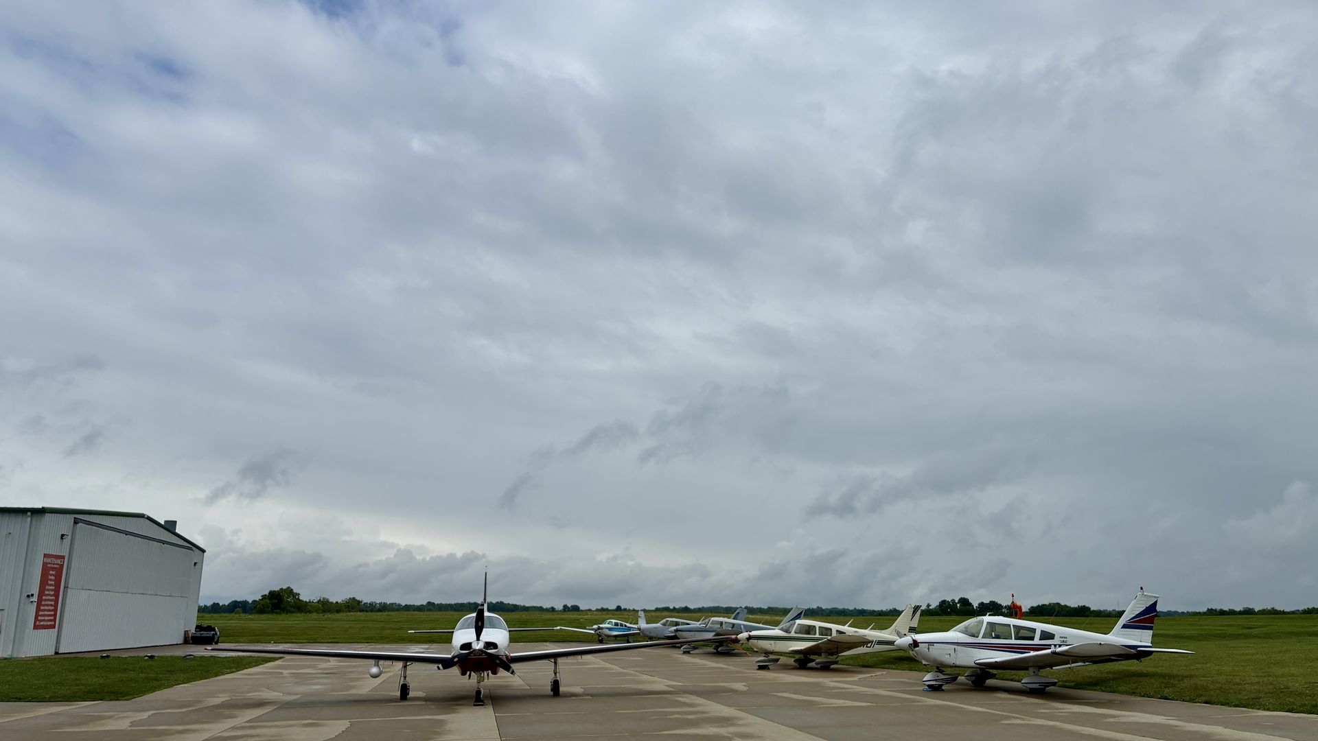 Overcast sky above a small airport. A single propeller plane faces the camera on a taxiway, with several small planes parked to the right and a hangar on the left.