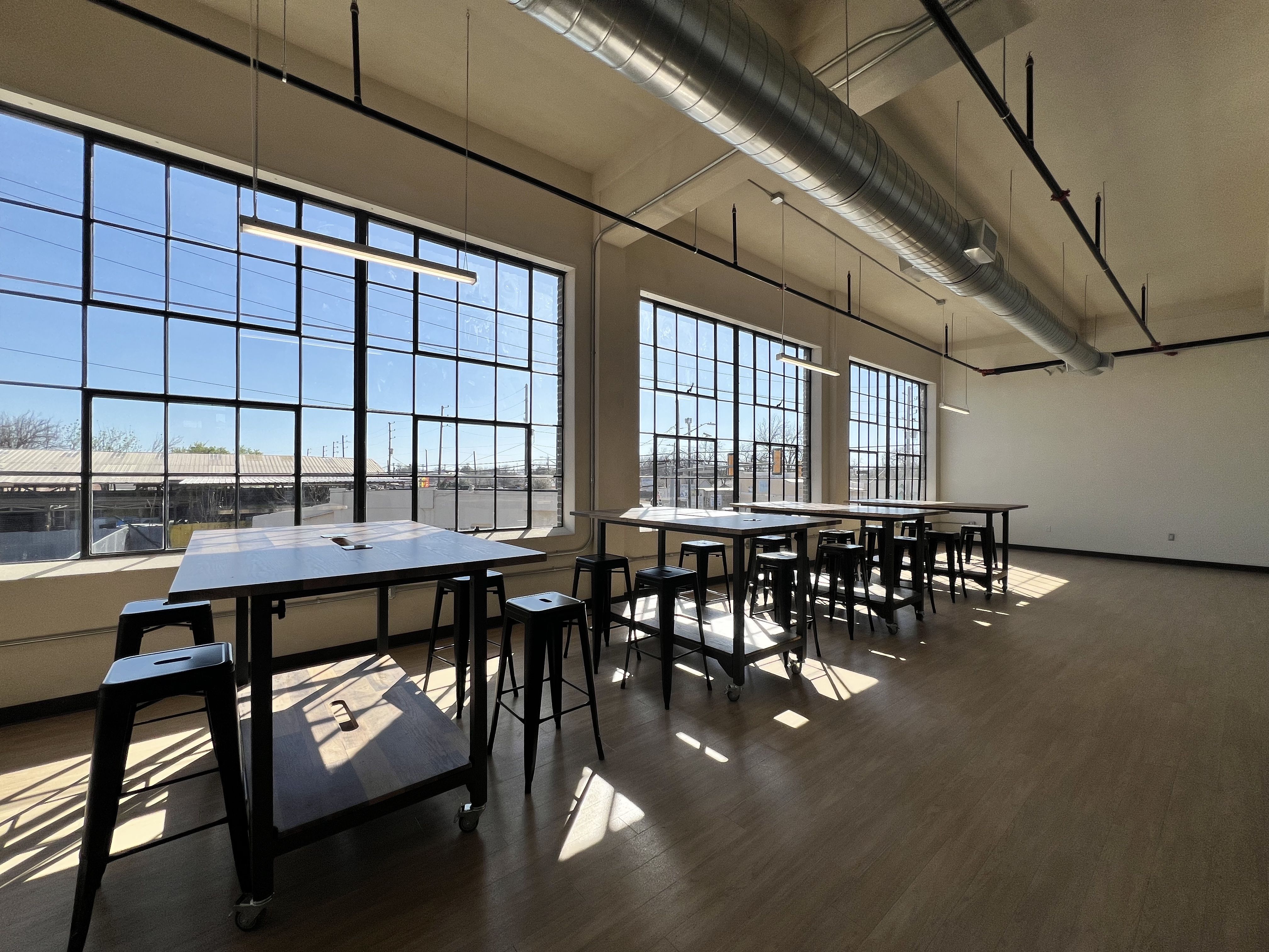 Bright industrial classroom with large grid windows along one wall, long white work tables on wheels, and black stools. Sunlight streams in, casting shadows; exposed ductwork runs across the ceiling.