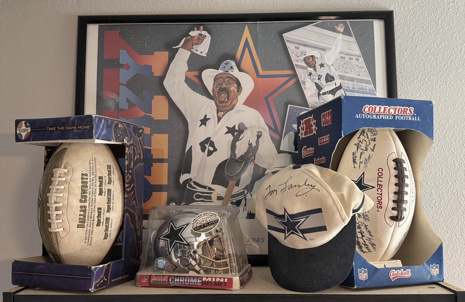 Dallas Cowboys memorabilia including two boxed footballs, a 24k gold-plated mini helmet, a signed cap, displayed in front of a framed colorful poster featuring a man in a cowboy outfit.