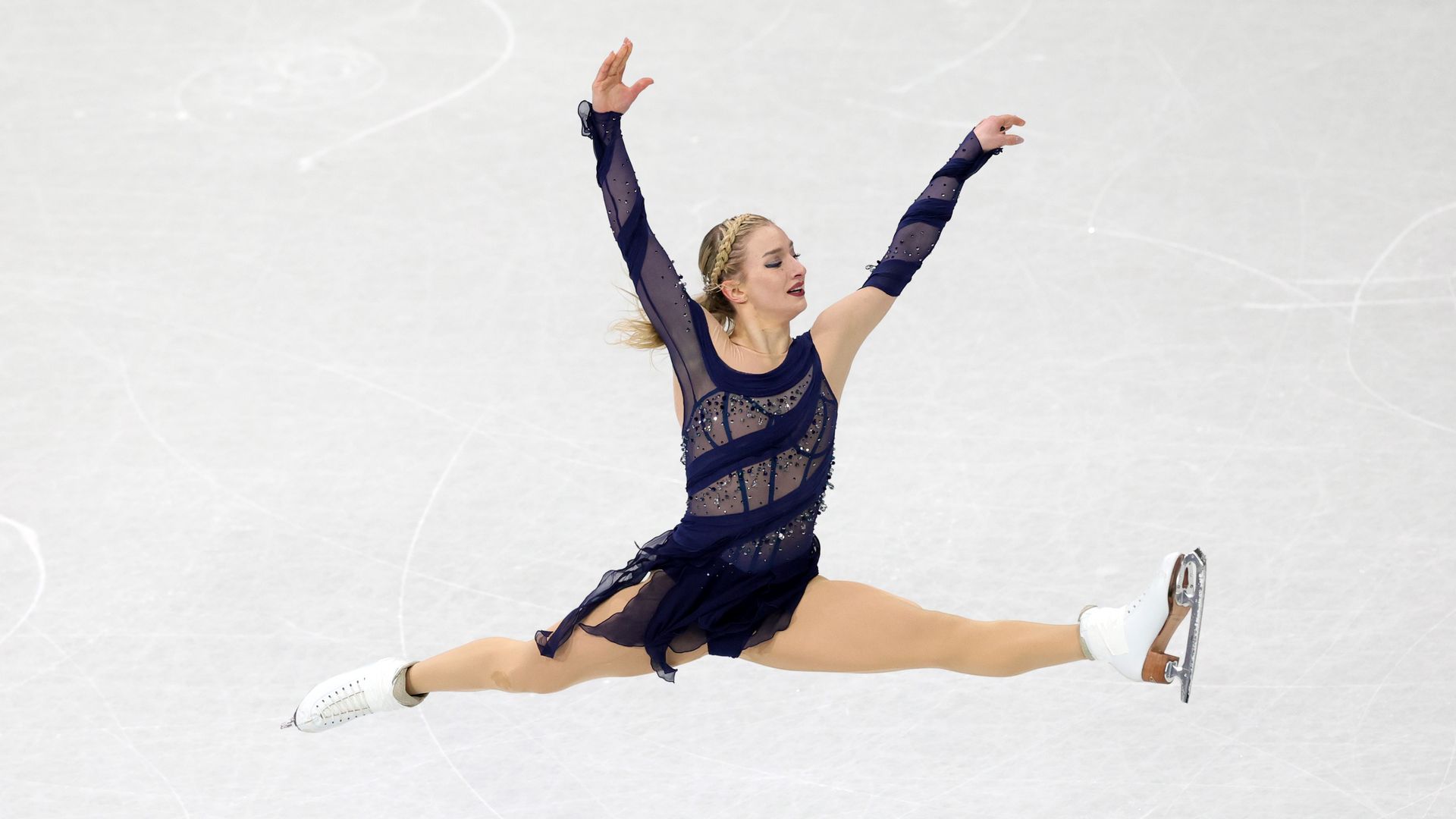 Amber Glenn, wearing a navy blue figure skating outfit, hovers above the ice in a split jump at the 2026 Winter Olympics. 
