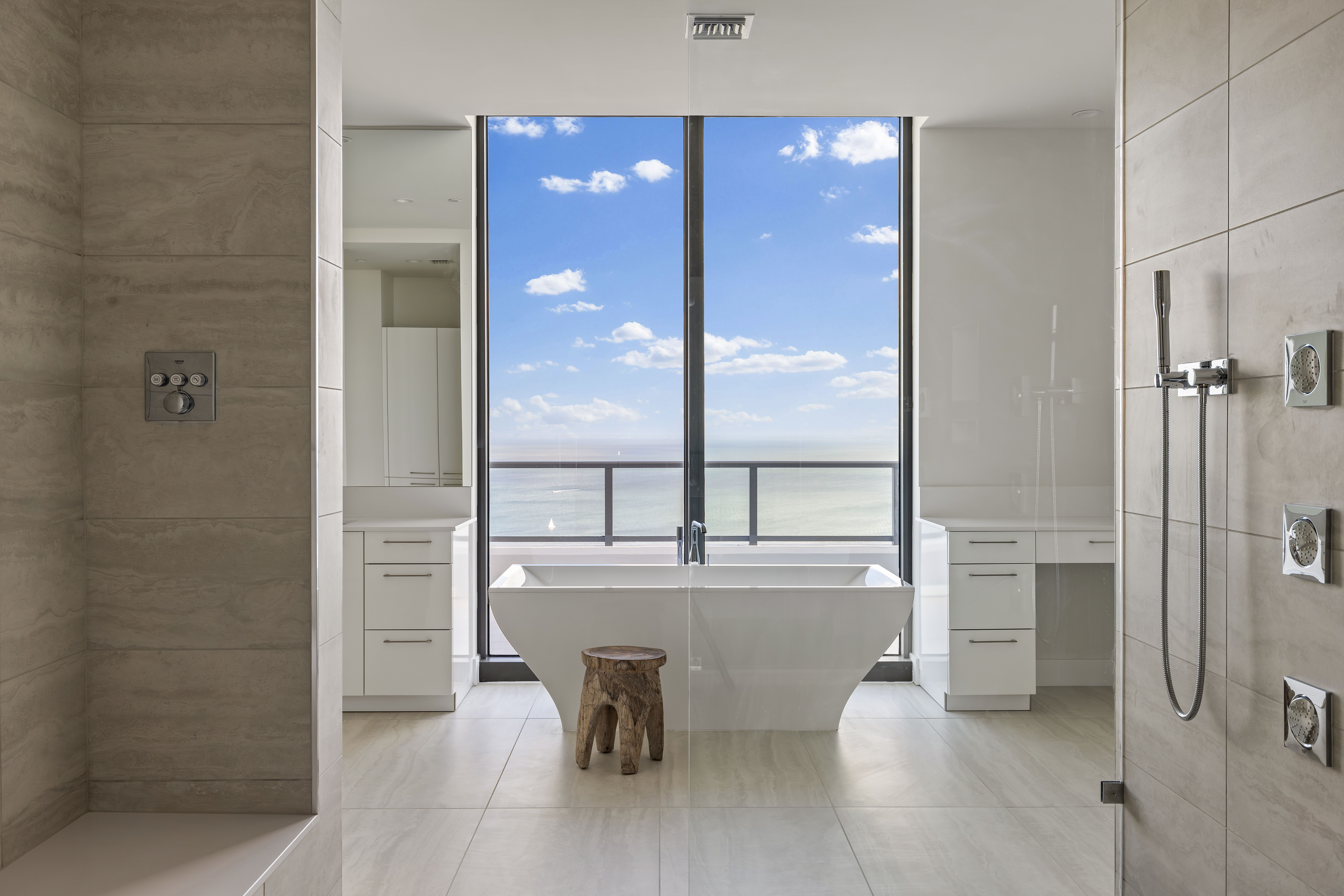 A bathroom with white cabinetry and sand-colored tiles, with the focal point a deep white bathtub positioned in front of two floor-to-ceiling windows with water views.