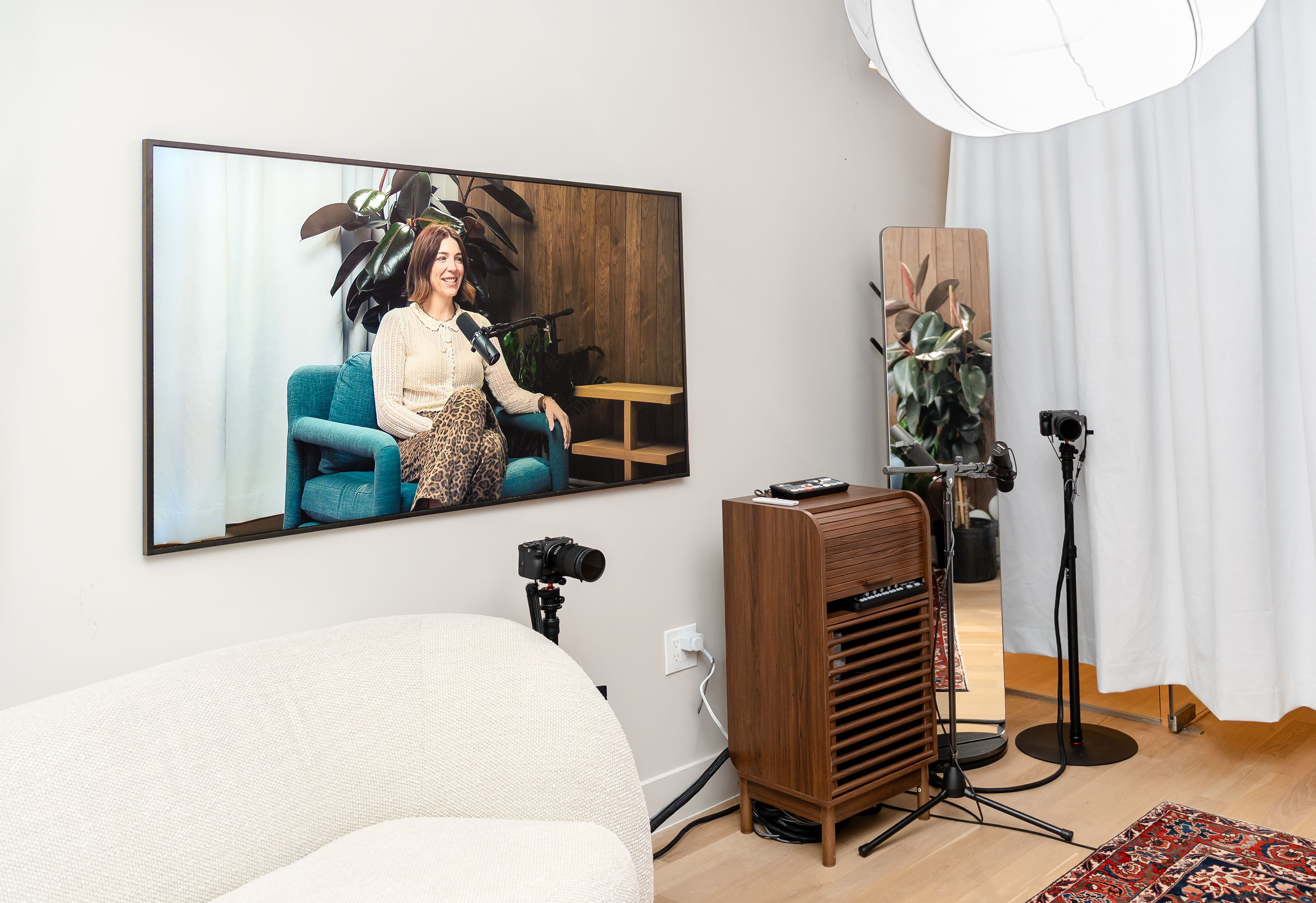 Person seated in a teal chair in a home recording studio, smiling into a microphone. A camera on a tripod and wooden cabinet sit near a large wall mirror reflecting plants; gray walls and curtains.
