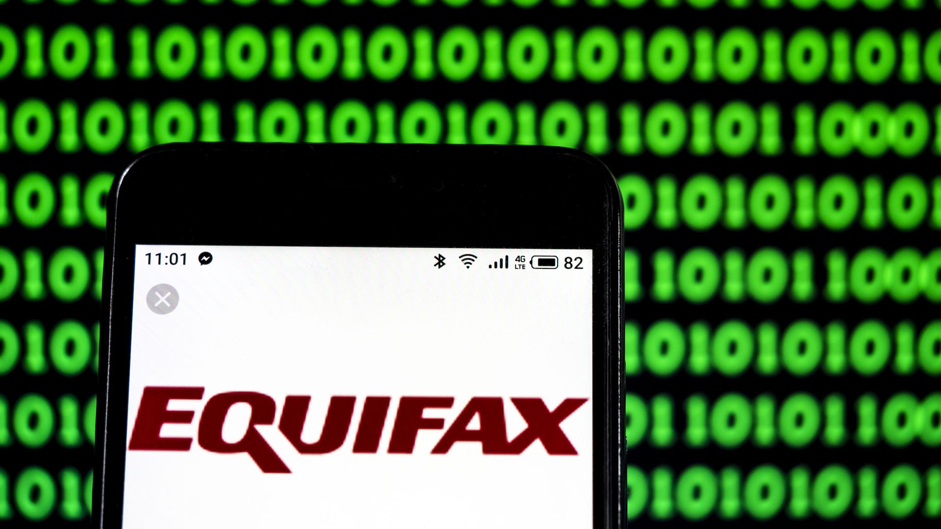 In this image, an iPhone is held close to the viewer against a green and black background of 1s and 0s. The phone screen is a white background with the logo 'Equifax' displayed in red.