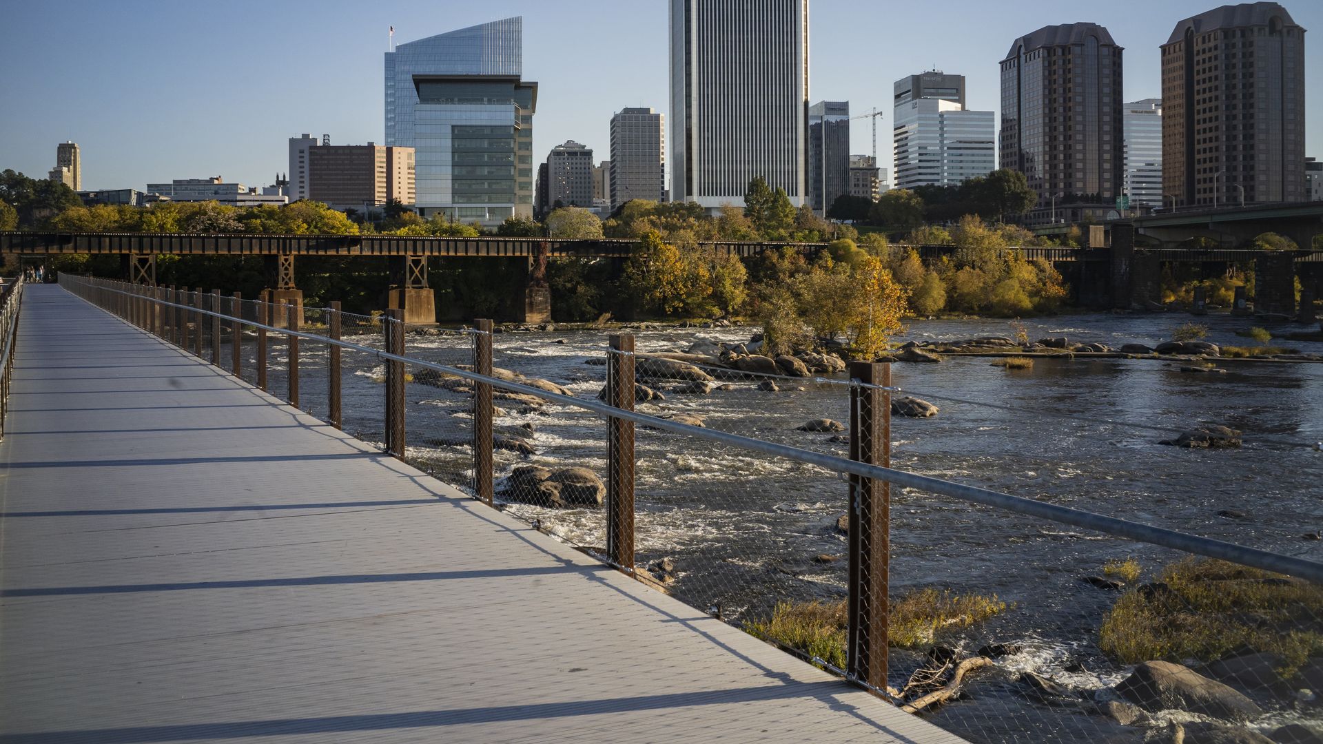 Sunlit city skyline across a river with glass and concrete high-rises. A wide walkway with a chain-link railing runs along the water, autumn trees glow in gold and green under a blue sky.
